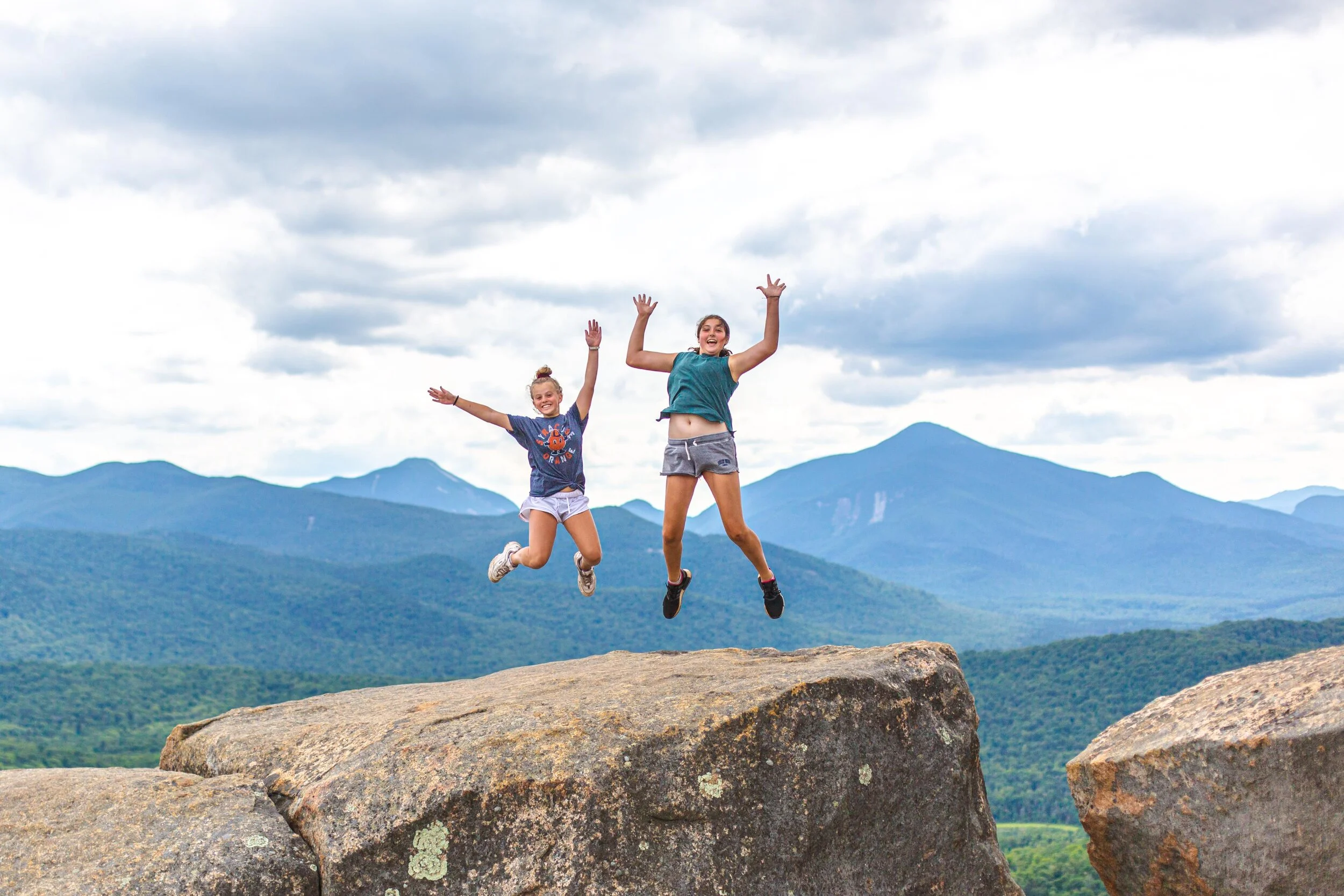 Two girls jumping off large rocks with a mountain landscape and cloudy sky in the background