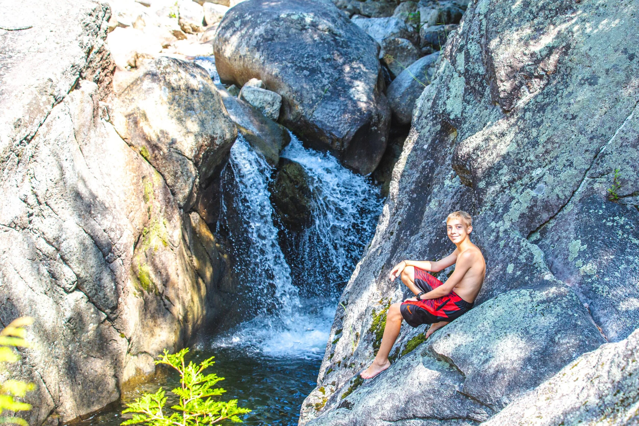 A young boy in red and black shorts sitting on a large rock beside a small waterfall in a rocky creek with greenery nearby.