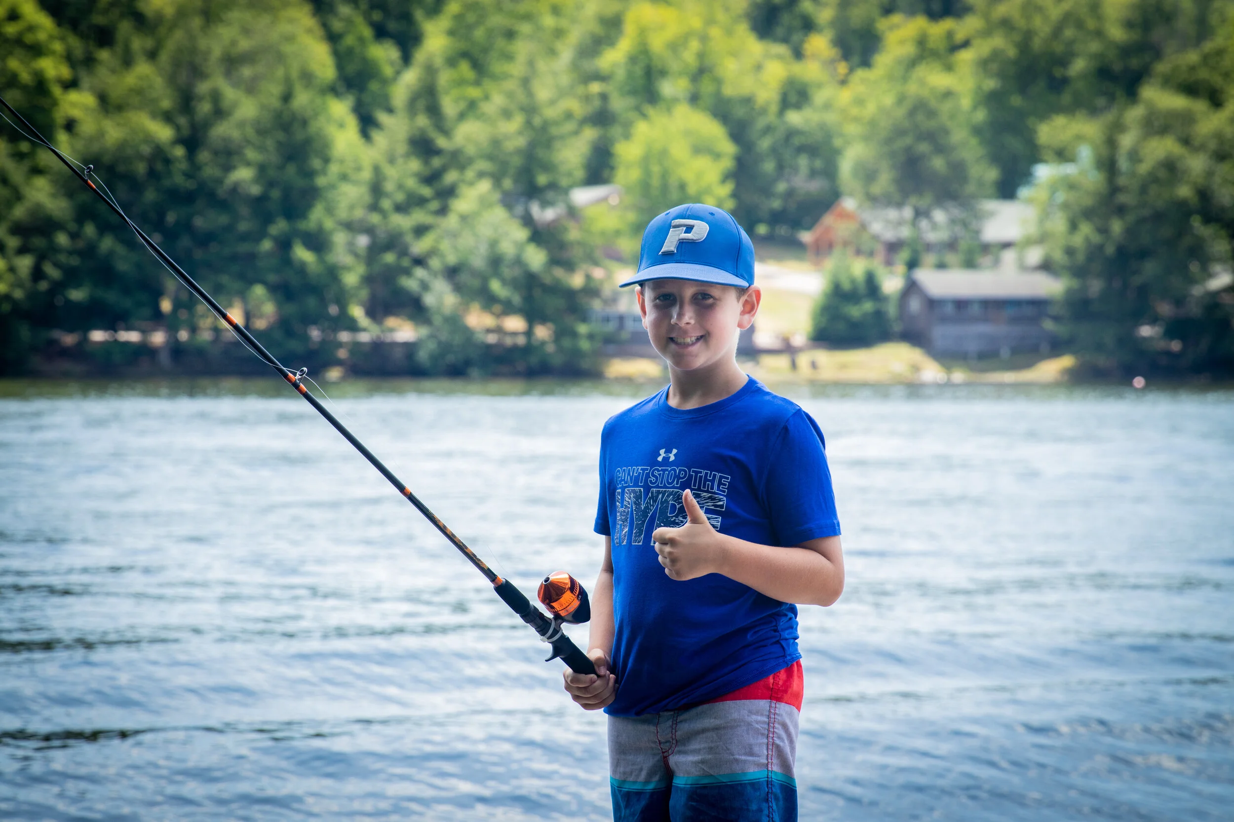 Young boy fishing by a lake, smiling and giving a thumbs up while holding a fishing rod, with green trees and houses in the background.