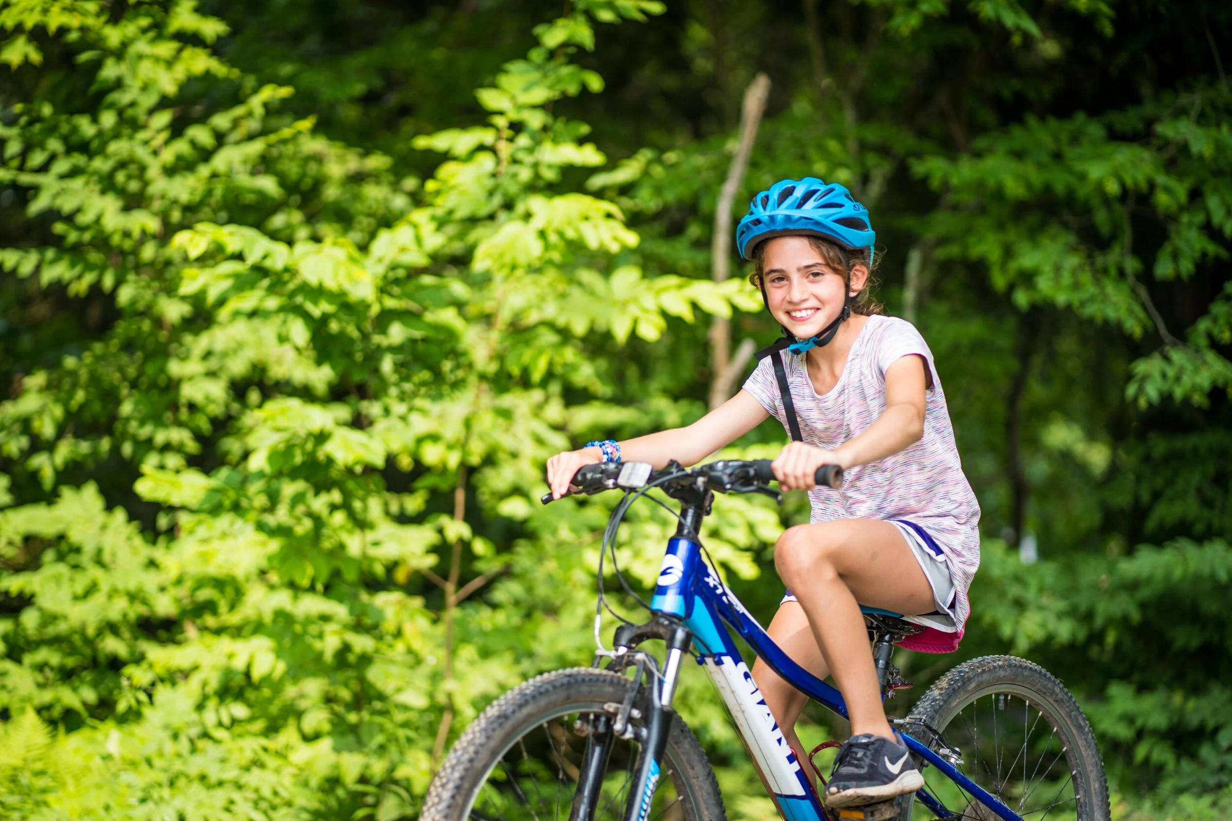 Young girl riding a blue mountain bike in a green forest, wearing a blue helmet and smiling.