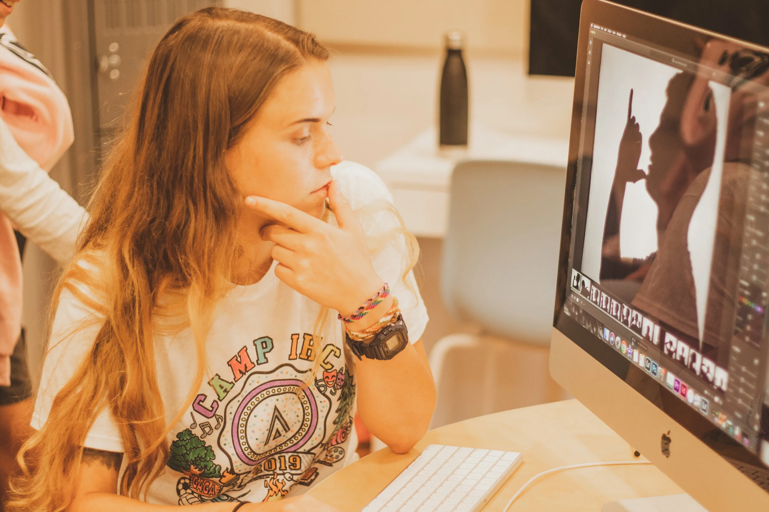 A young woman with long wavy hair, wearing a white t-shirt with colorful text and graphics, sitting at a desk looking at a computer screen with concentrated interest, hand on chin, in an indoor setting.
