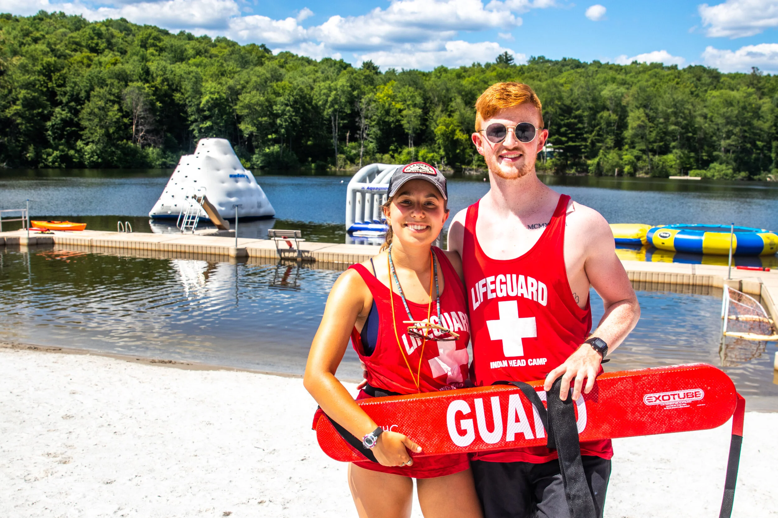 Two lifeguards, a woman and a man, standing on a beach near a lake, both wearing red lifeguard shirts, smiling and holding a rescue board. In the background, there's a small pool, a water rescue training float, and a floating climbing structure on th