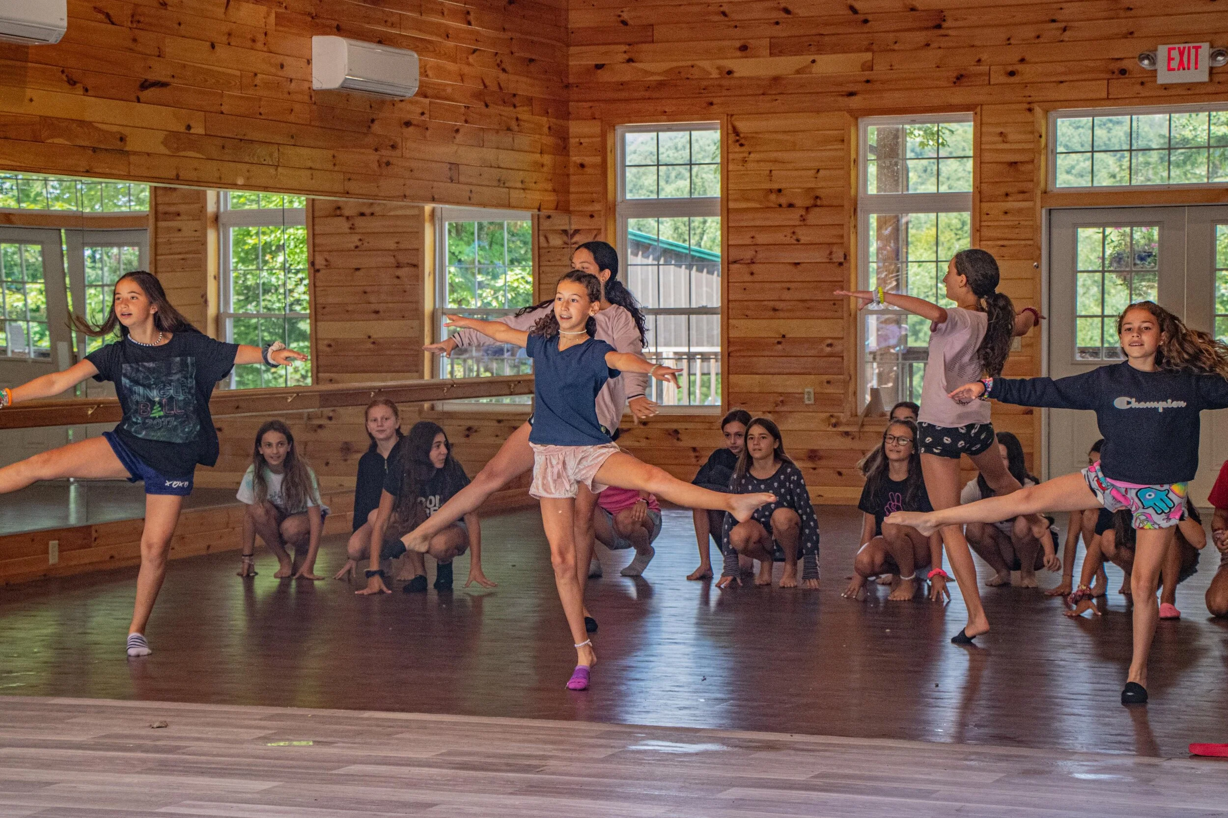 Young girls practicing ballet or dance moves in a wooden-finished dance studio with large windows and a group of children watching.