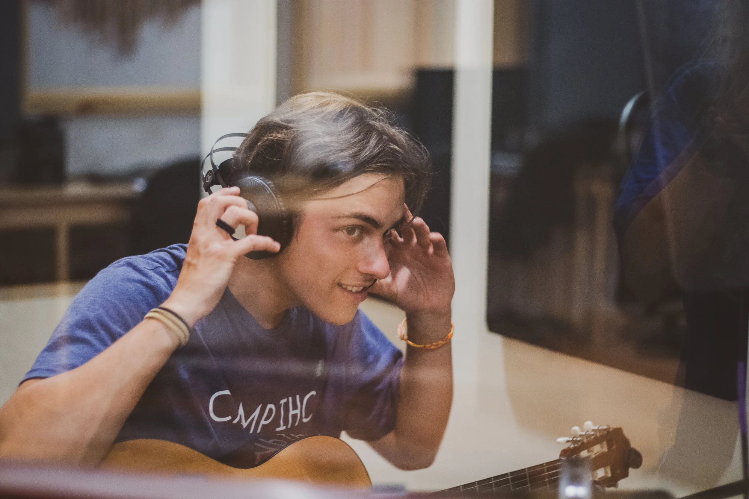 A young person smiling while looking through a glass at a recording studio, holding headphones to their ears and playing an acoustic guitar.