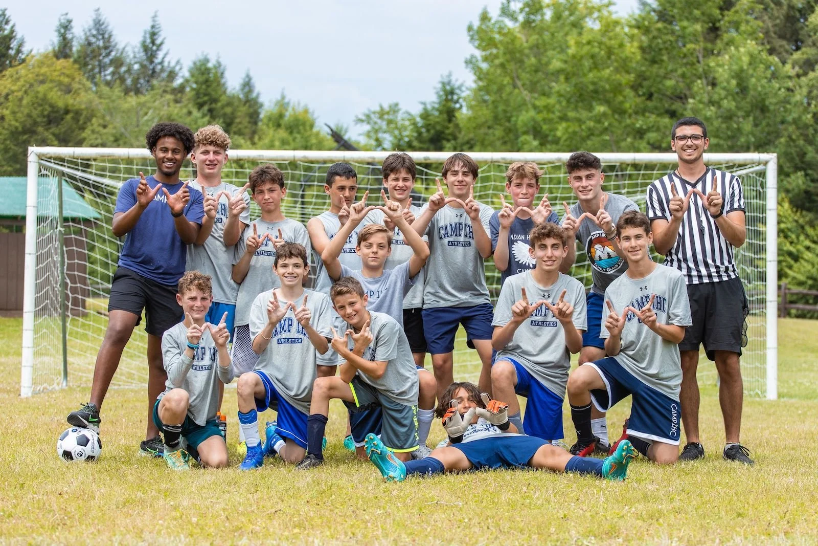 Group of young boys and their coaches on a soccer field posing for a team photo in front of a goal, wearing sports attire, some making hand gestures, with a soccer ball on the grass.