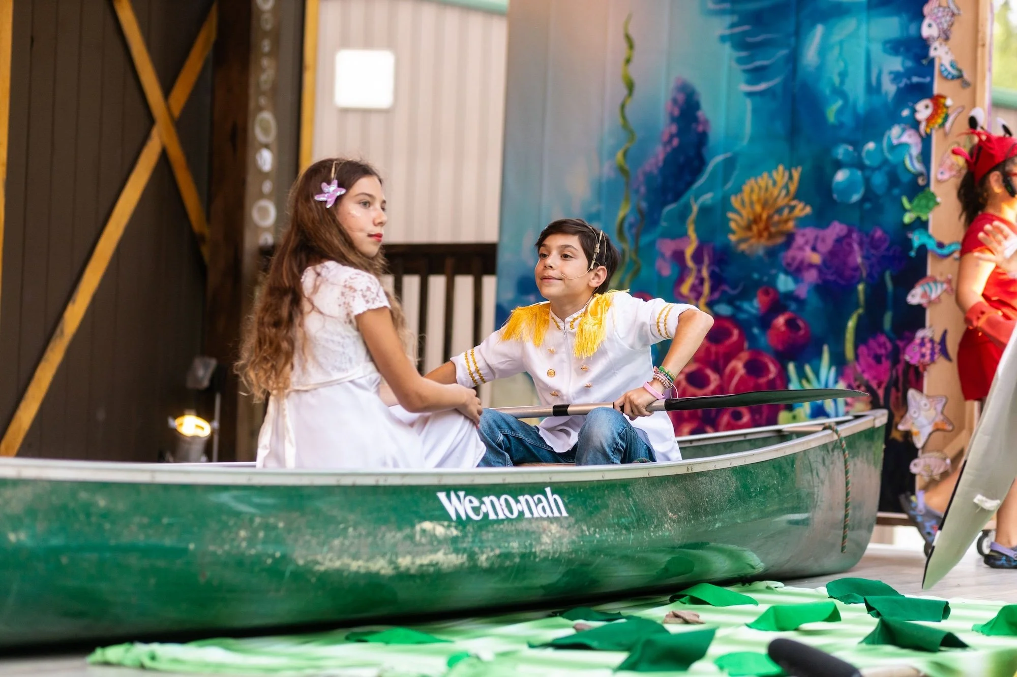 Two children dressed as characters, a girl in a white dress and a boy in a white shirt with yellow fringe, in a green boat with the word "We-no-nah" on the side, part of a stage set with an underwater scene backdrop and other children in costumes nea