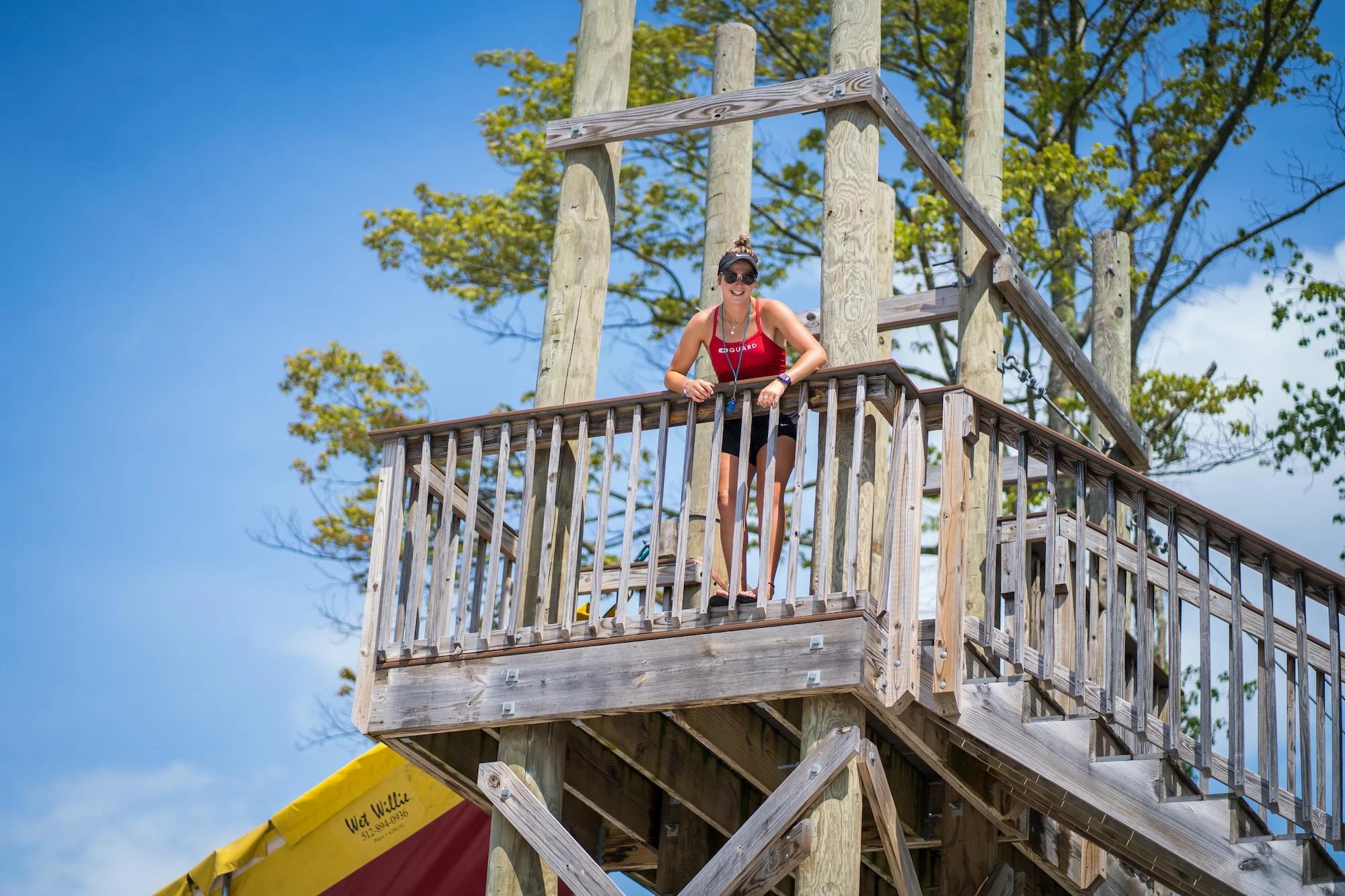 A woman standing on a wooden platform at a treehouse, smiling, with a blue sky and trees in the background.