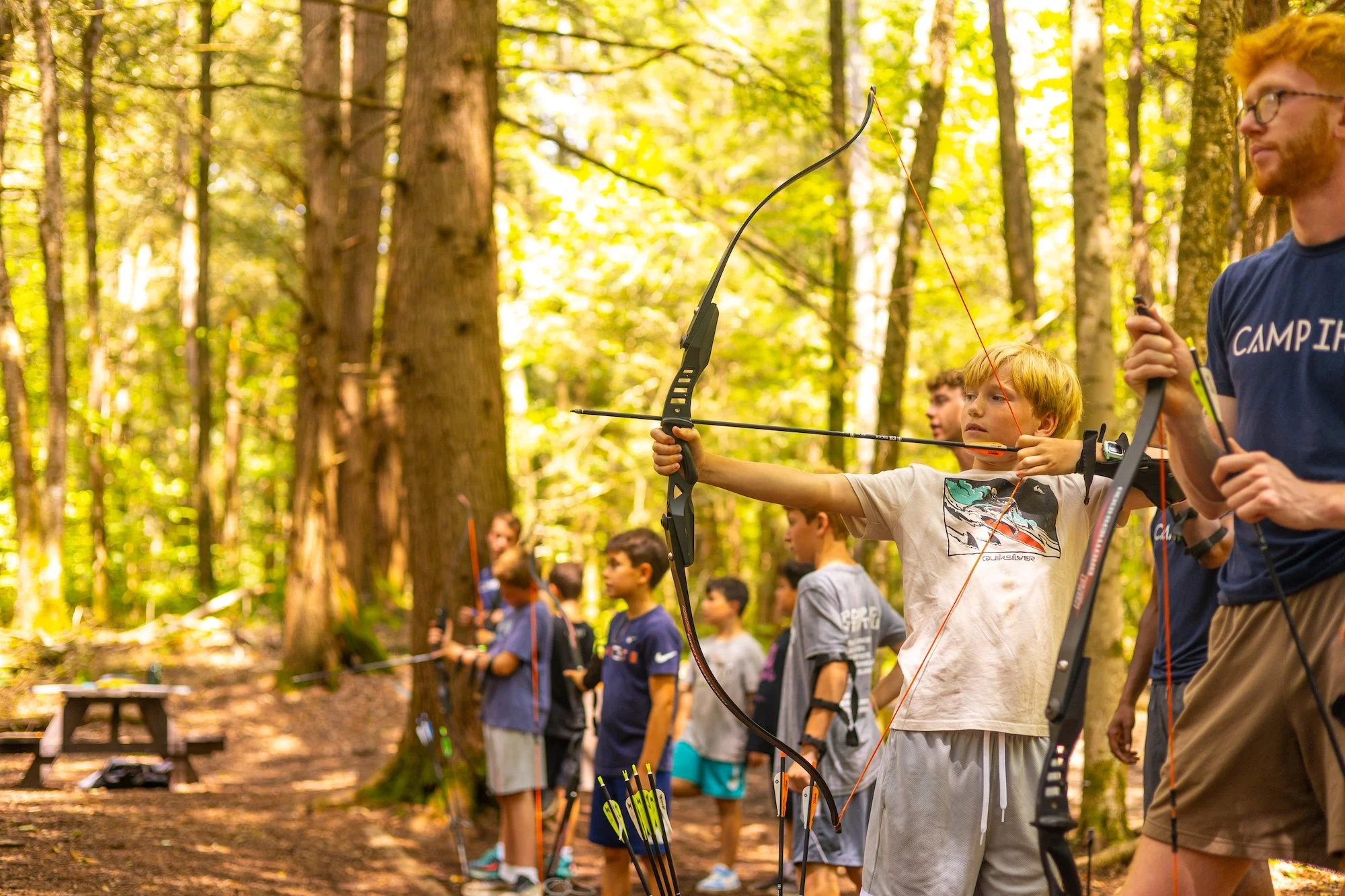 Children and an adult outdoors in a forest, participating in archery with bows and arrows.
