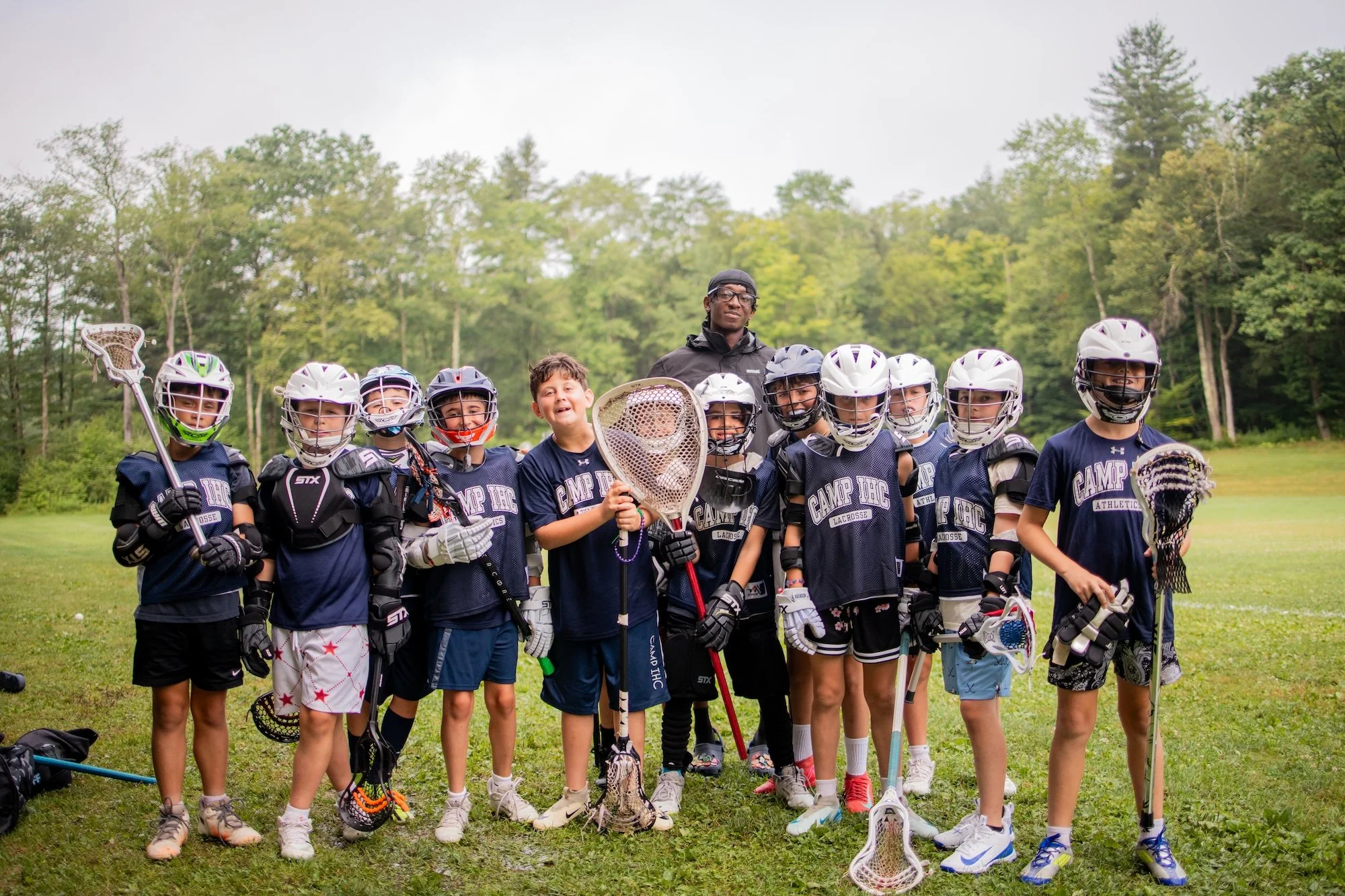 Group of boys and a coach standing outdoors on grass, wearing lacrosse gear and jerseys, holding lacrosse sticks, with trees in the background.