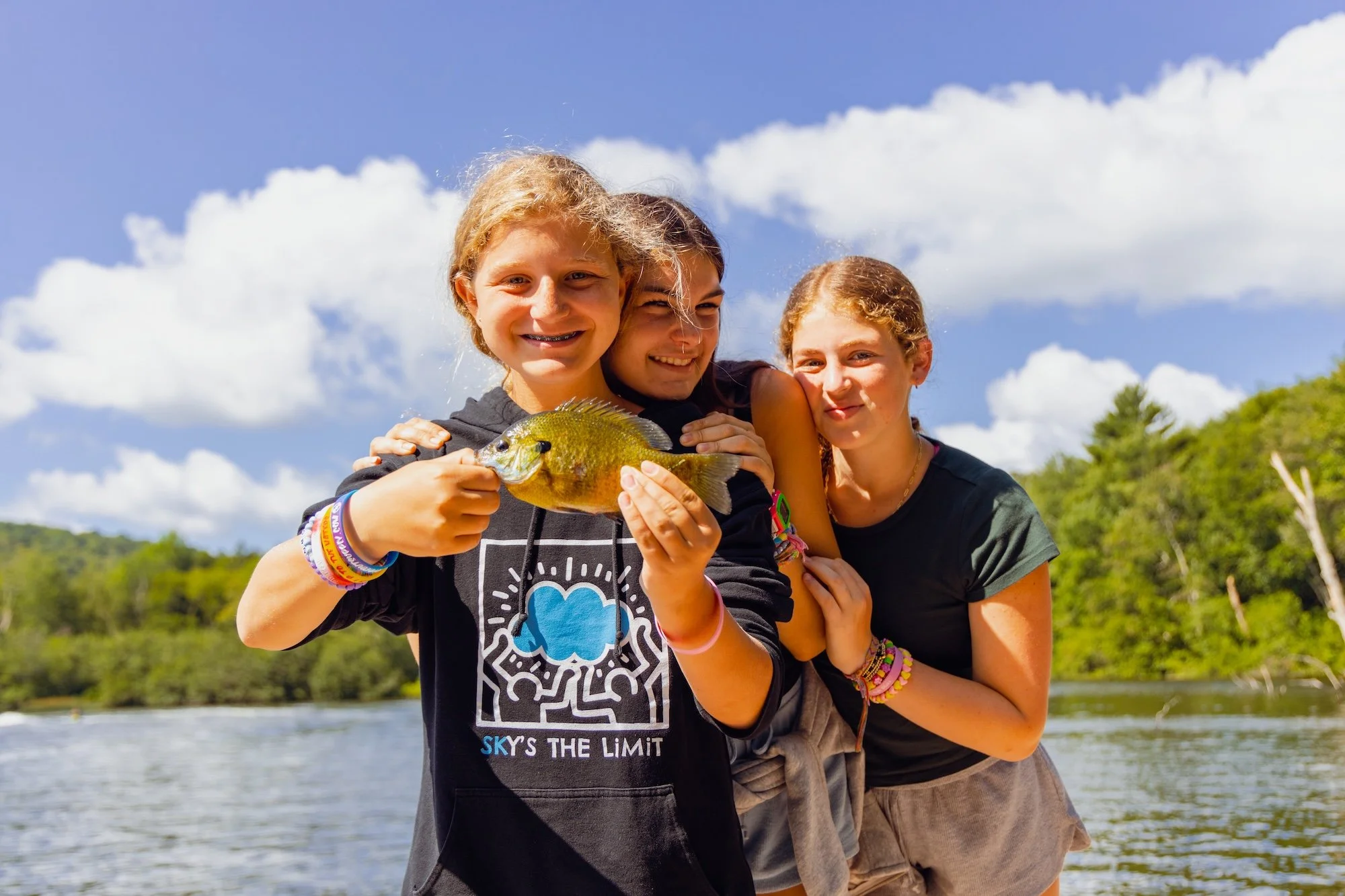 Three girls smiling on a boat holding a fish with a green landscape and blue sky in background.