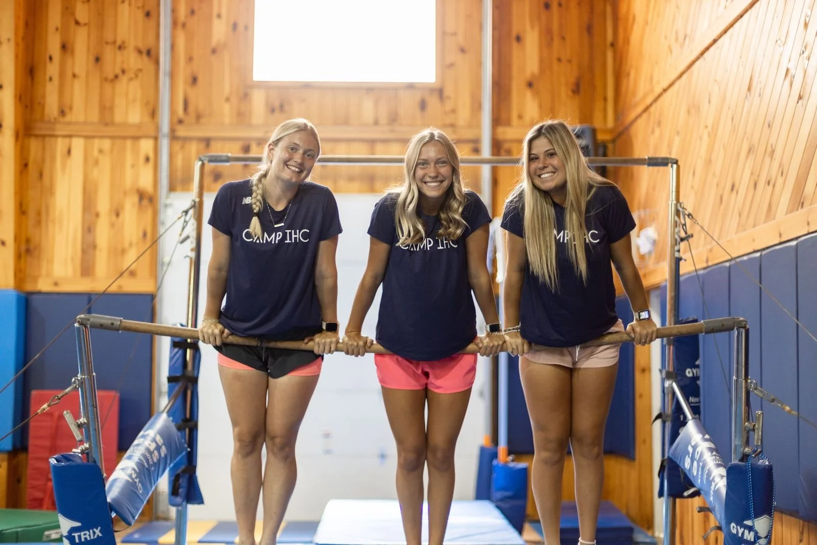 Three young women in navy t-shirts and shorts smiling and holding onto a gymnastics balance beam in a wood-paneled gymnasium.