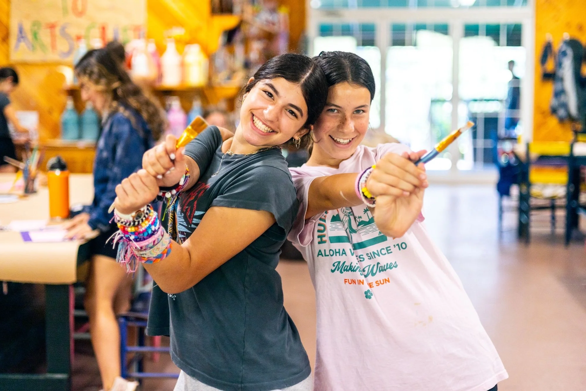 Two smiling teenage girls standing back-to-back inside a craft or art store holding paintbrushes, teaching or creating art together, with art supplies visible in the background.