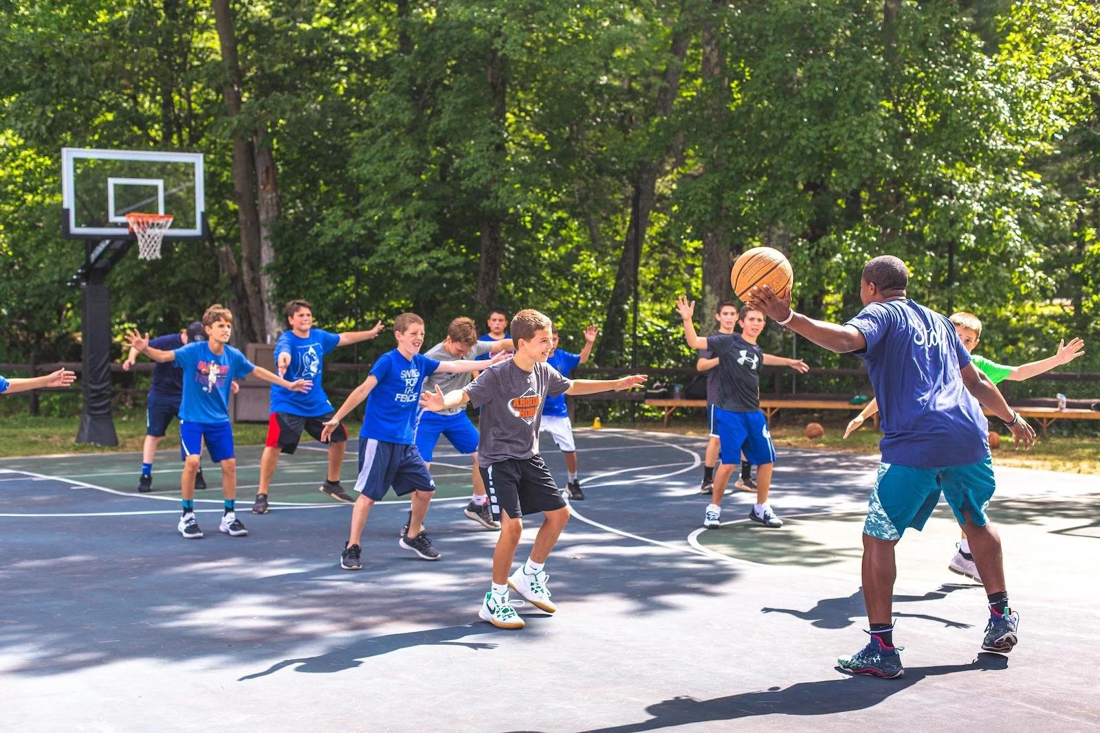 A basketball coach instructs a group of young boys on an outdoor basketball court during daytime, with the boys practicing defensive stance and wearing casual athletic clothing, surrounded by trees.