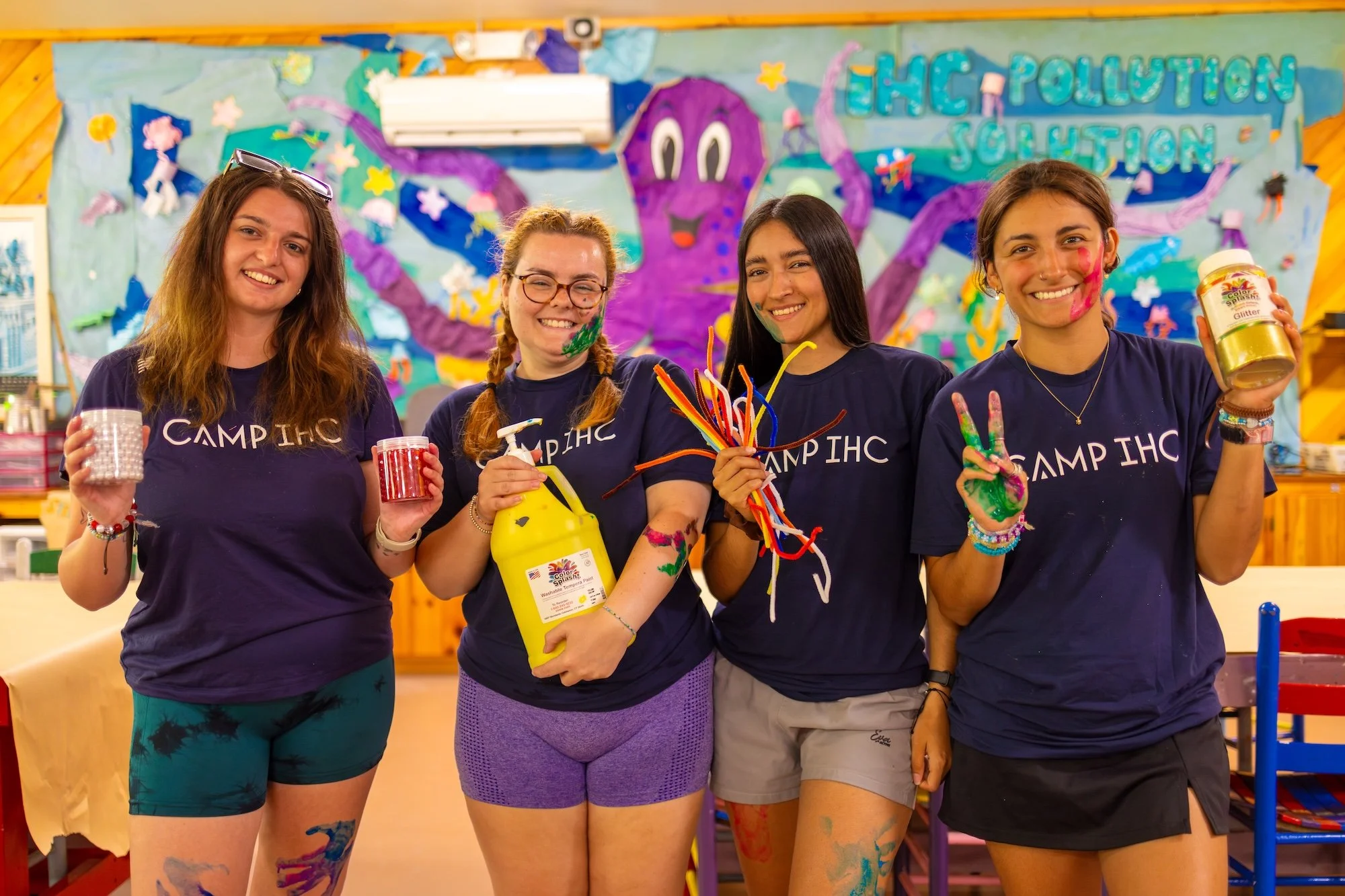 Four young women smiling and holding art supplies in front of an underwater-themed decoration with a purple octopus and the words 'EHC Pollution Solution'.