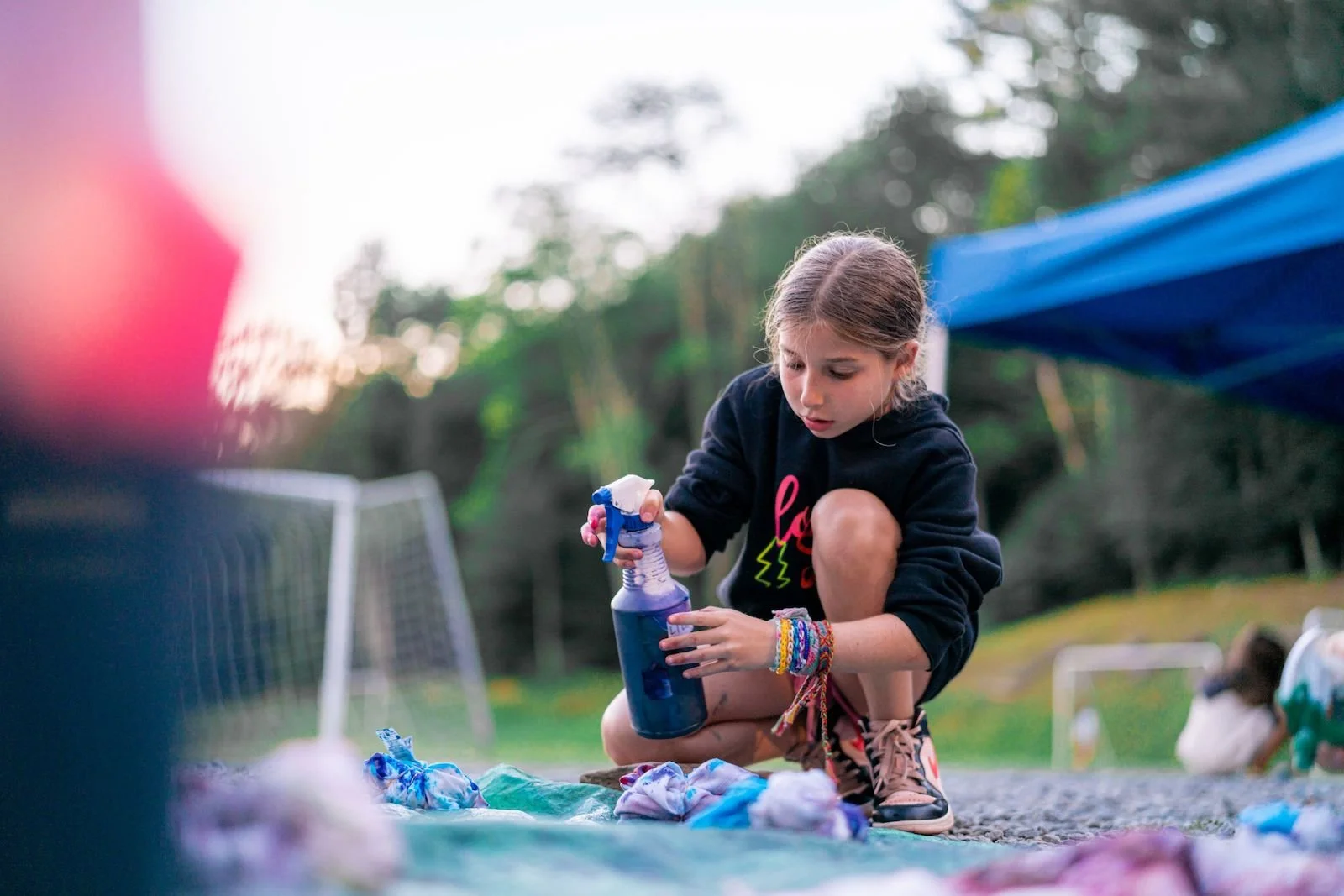 A young girl crouches outdoors, spray cleaning a piece of cloth or clothing with a spray bottle during daytime, with blurred trees and other children in the background.