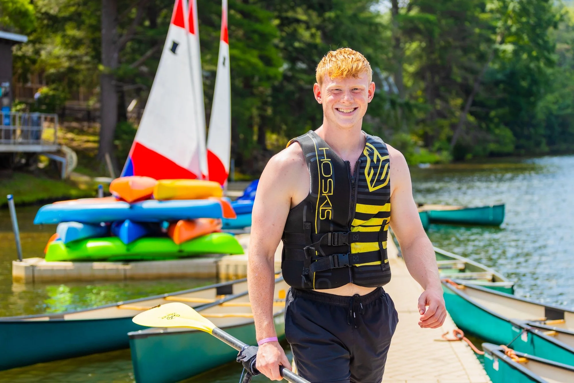 A young man smiling on a dock with kayaks and sailboats, holding a paddle, in a sunny outdoor setting with trees and water.