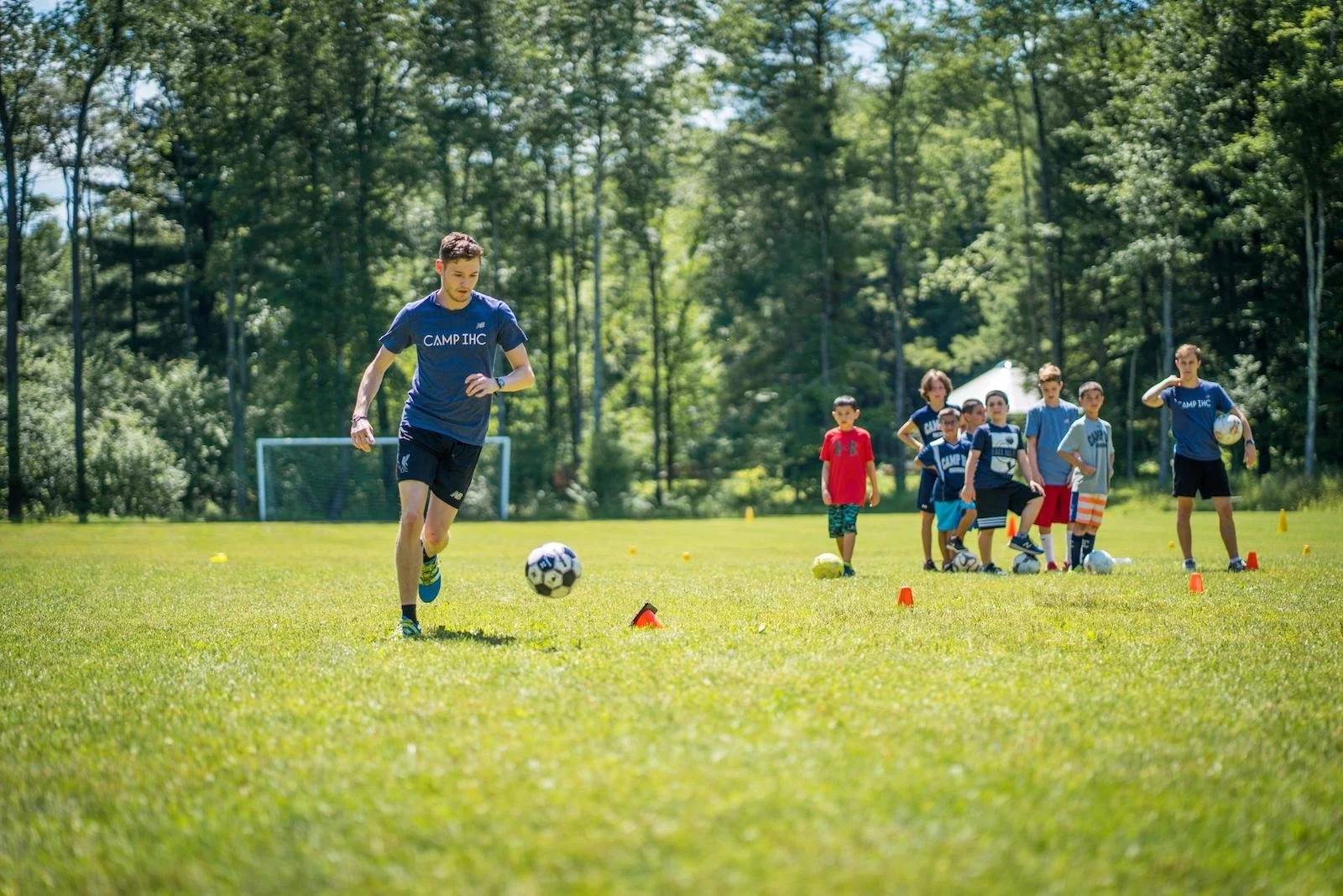 Young males playing soccer during a training session on a sunny field surrounded by trees, with cones set up for drills.