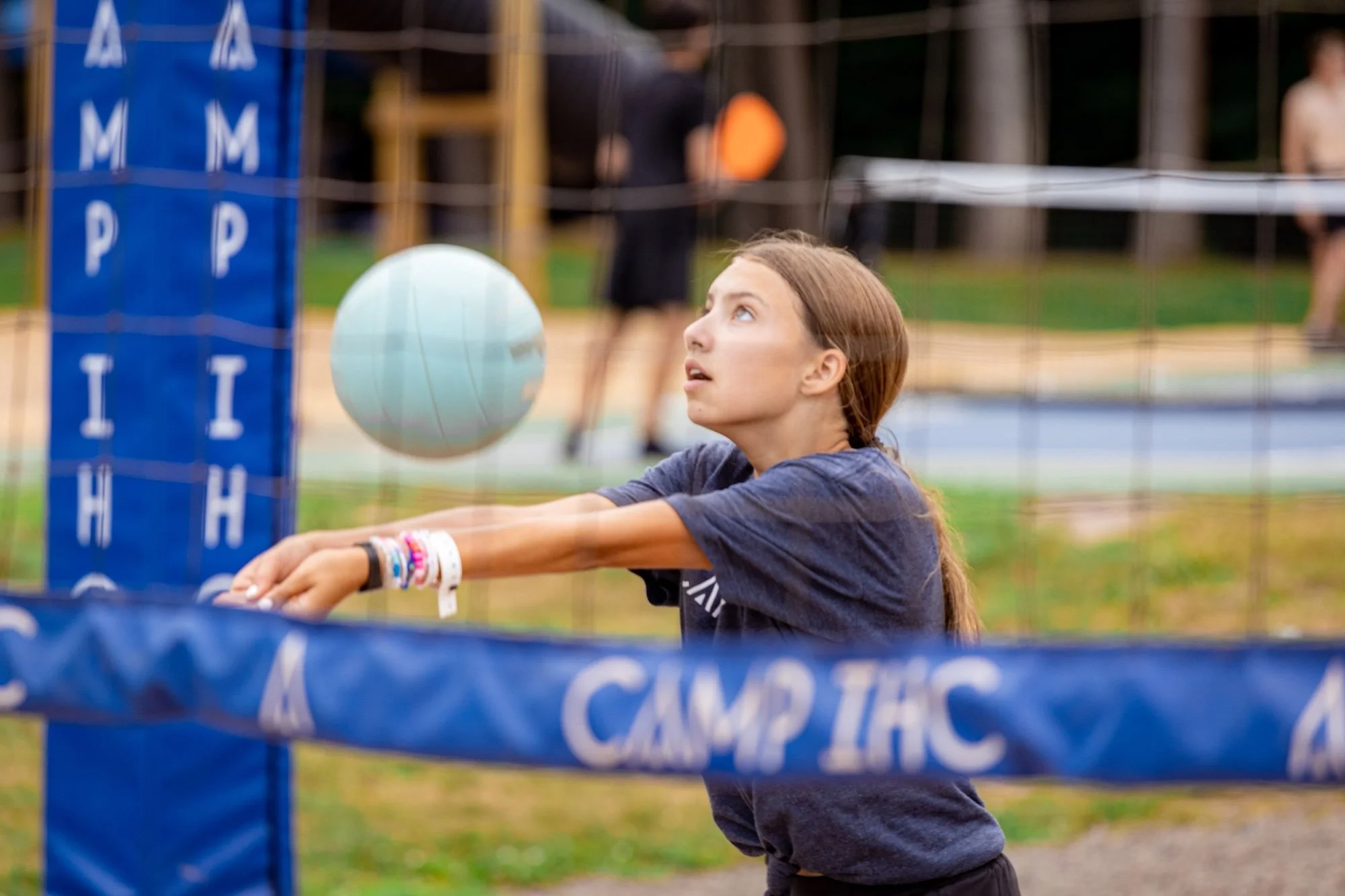 Young girl reaching out to hit a volleyball at an outdoor volleyball court with a net marked 'CAMP IAC' in the foreground.
