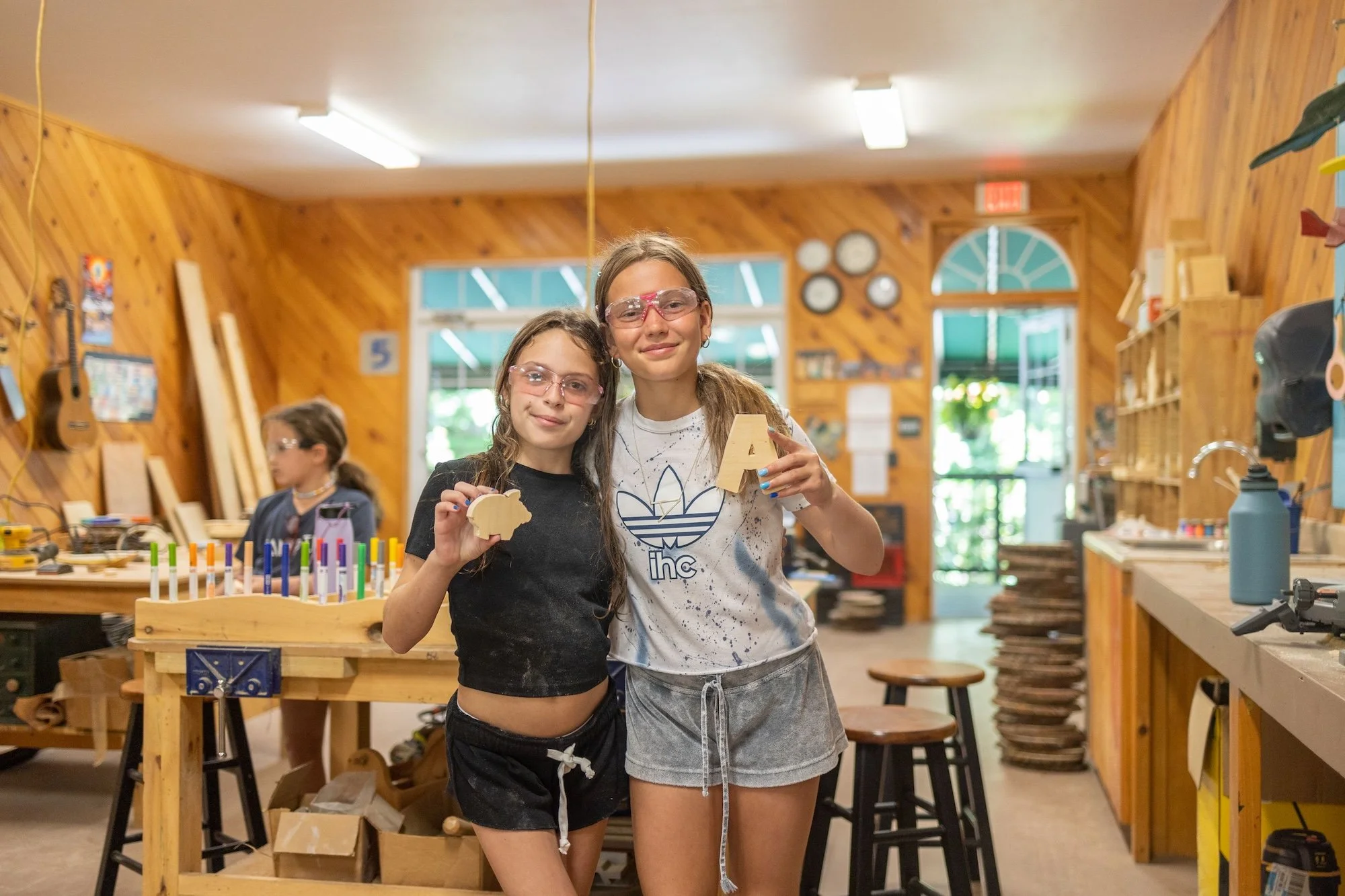 Two young girls wearing safety glasses standing in a woodworking shop, holding small wooden objects and smiling at the camera.