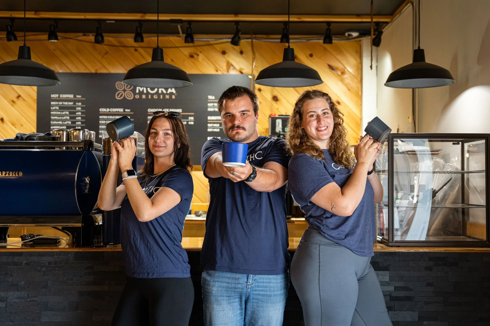 Three baristas in navy blue shirts standing behind a coffee bar, each holding coffee mugs, smiling and posing for the camera in a coffee shop with modern industrial design.