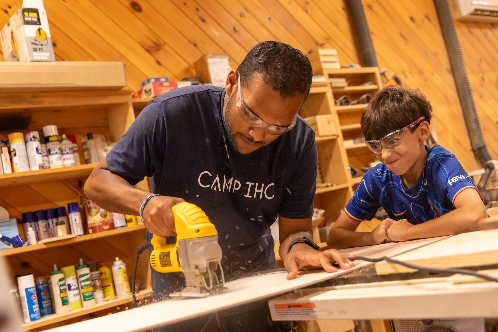 An adult and a young boy working together at a woodworking workshop, using a power saw on a piece of wood, both wearing safety goggles.