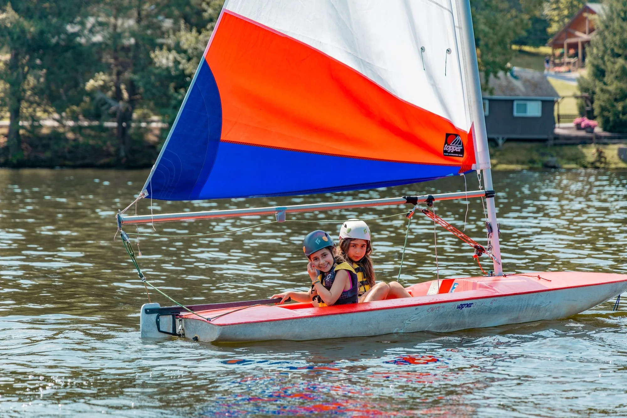 Two girls wearing safety helmets and life jackets sailing a small colorful sailboat on a lake with houses and trees in the background.