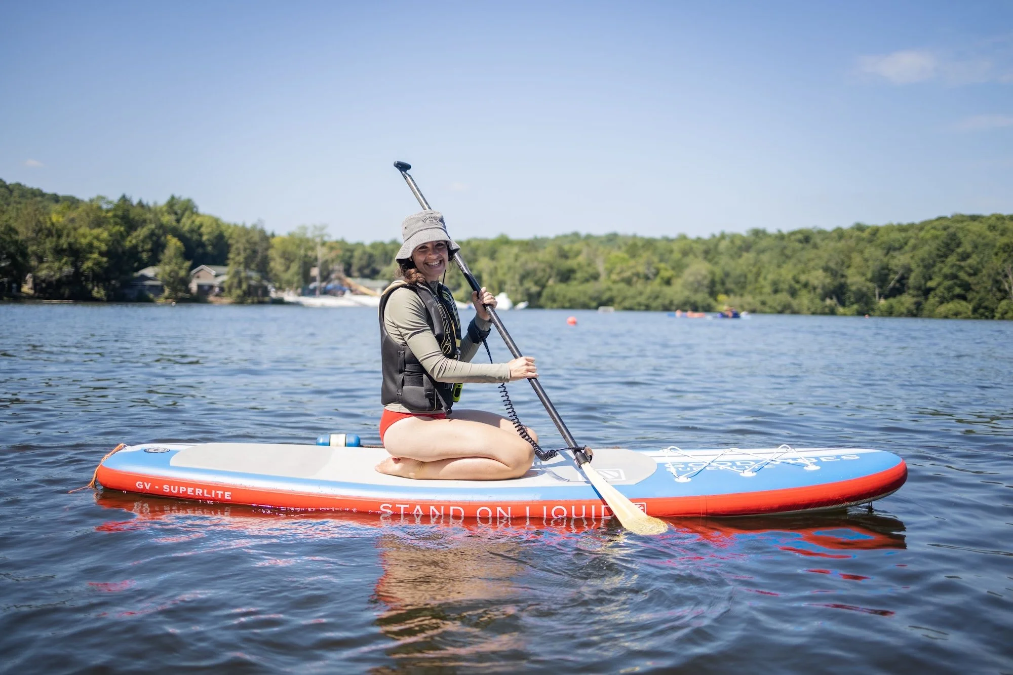 Woman kneeling on paddleboard on a lake, holding a paddle, wearing a hat, vest, and swimsuit, with trees and houses in the background under a blue sky.
