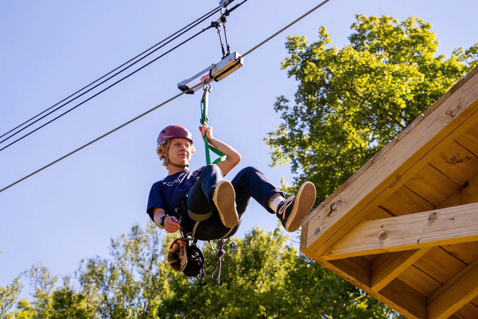 Young boy zip lining outdoors, wearing a red helmet and harness, jumping from a wooden platform, with green trees and blue sky in the background.