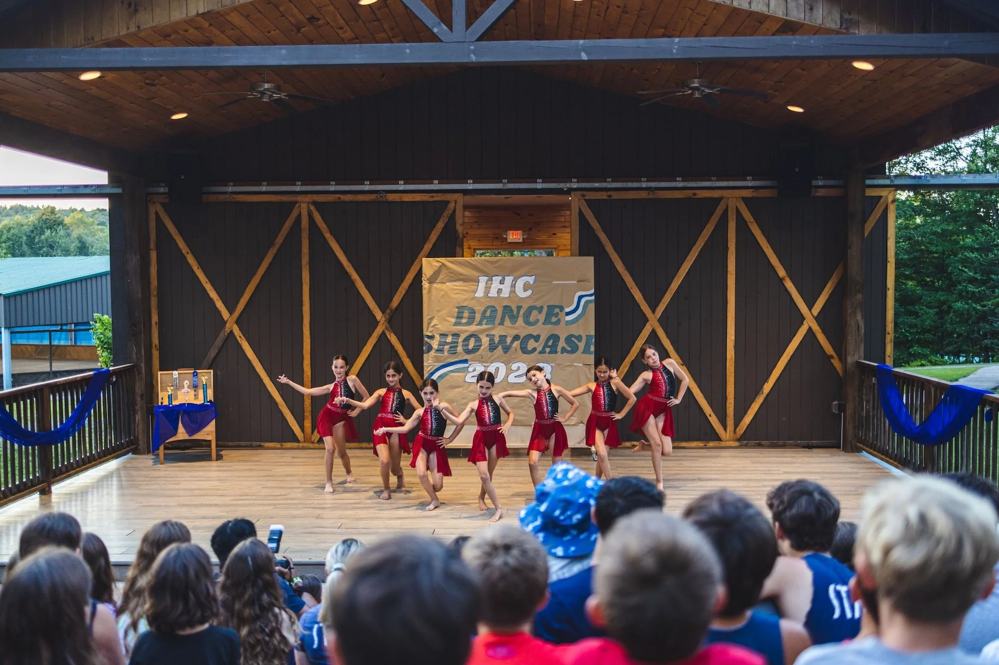 Group of young dancers performing on stage at the IHC Dance Showcase 2023, with an audience watching. The stage has a wooden backdrop with a yellow banner, and the dancers are wearing coordinated costumes.