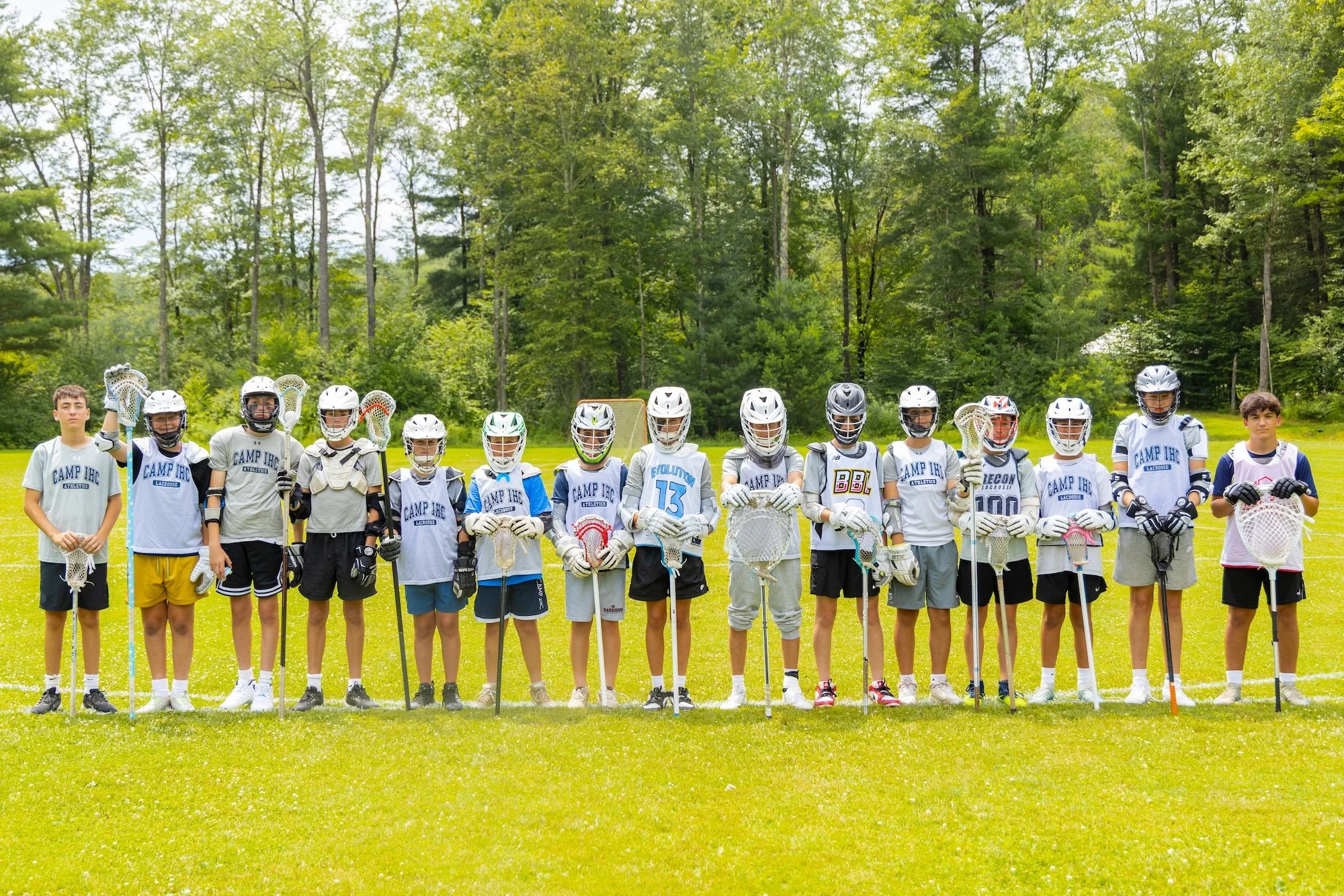 A young man and woman standing close together outdoors, smiling, with a group of people walking behind them in a wooded park.