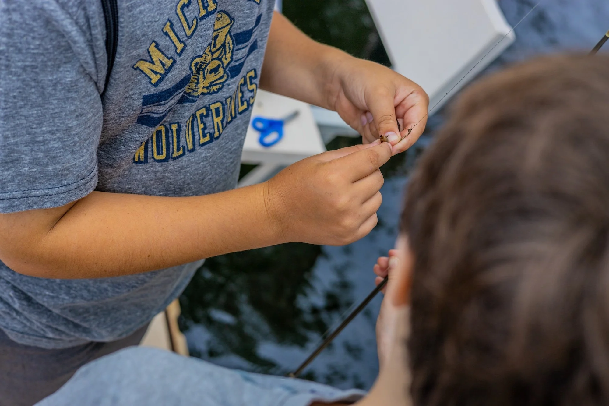 A person wearing a gray Michigan Wolverines shirt is holding a small fish and a fishing hook, while another person watches closely.