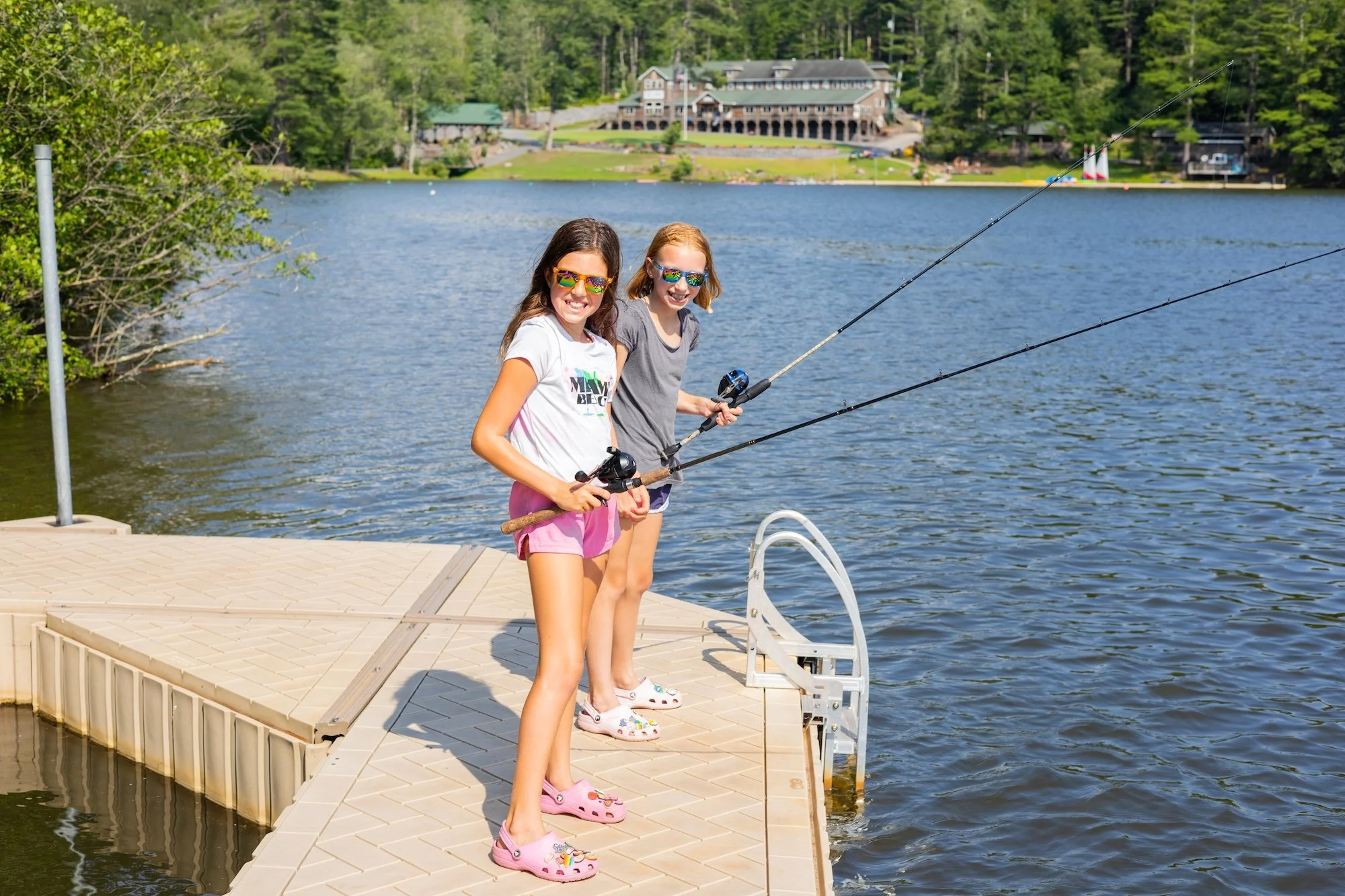 Two young girls are standing on a dock by a lake, fishing with rods. They are wearing colorful sneakers and sunglasses, smiling at the camera. In the background, there are trees and a large house by the water.