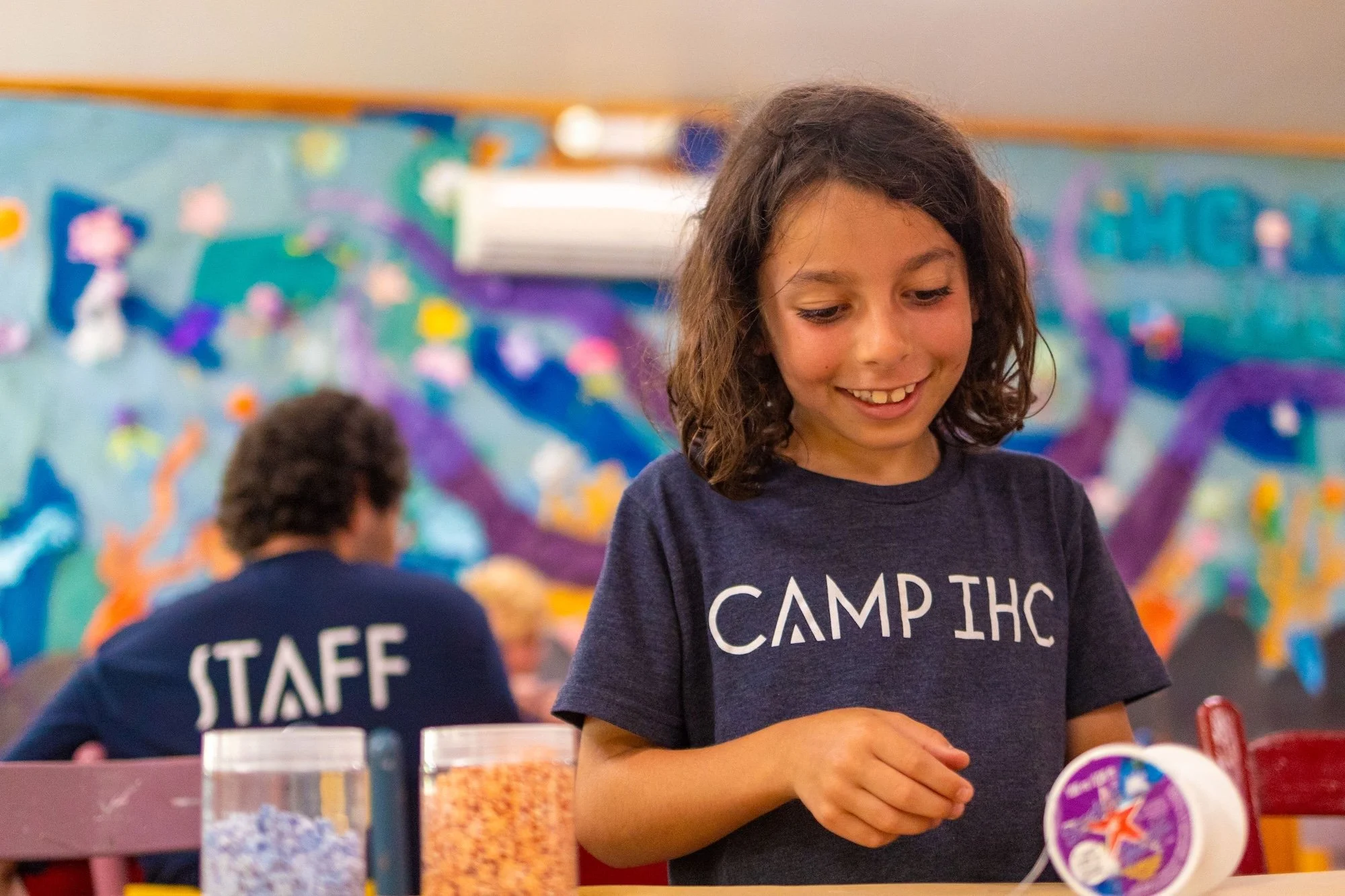A smiling young girl wearing a navy t-shirt with 'CAMP IHC' printed on it, working on a craft project at a table in an indoor activity space with colorful wall decorations. In the background, a person wearing a navy shirt with 'STAFF' on the back is
