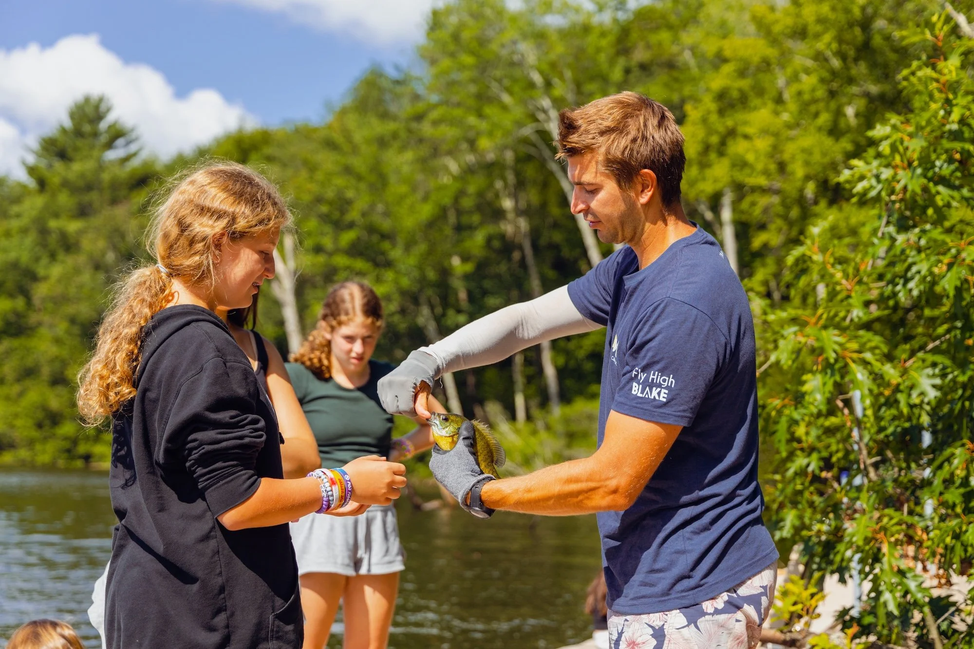 Guide shows a young girl a fish by a river with two other young girls watching, surrounded by green trees on a sunny day.