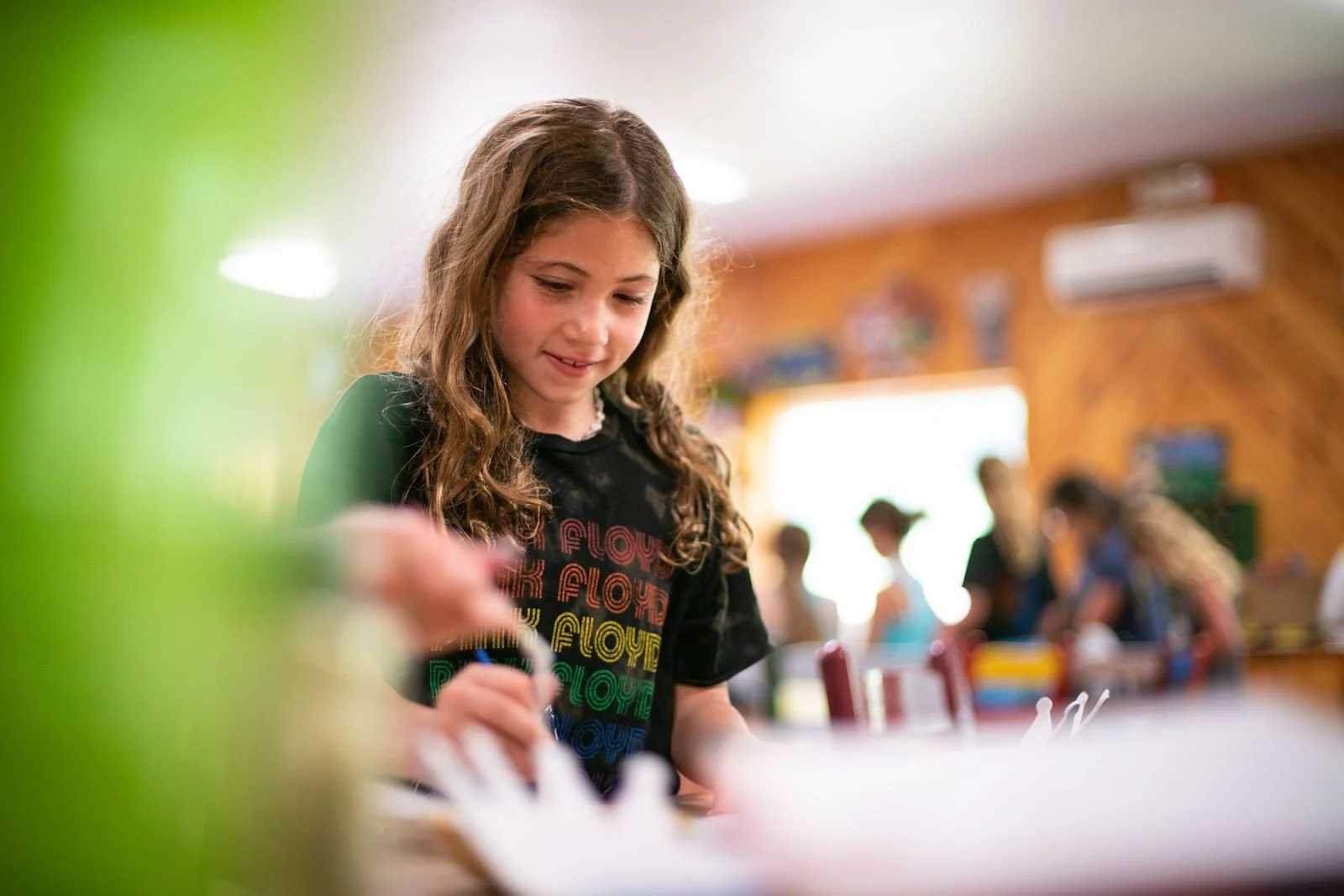 A young girl with long, curly brown hair working on a project in a classroom filled with other students. She is wearing a black t-shirt with colorful text.