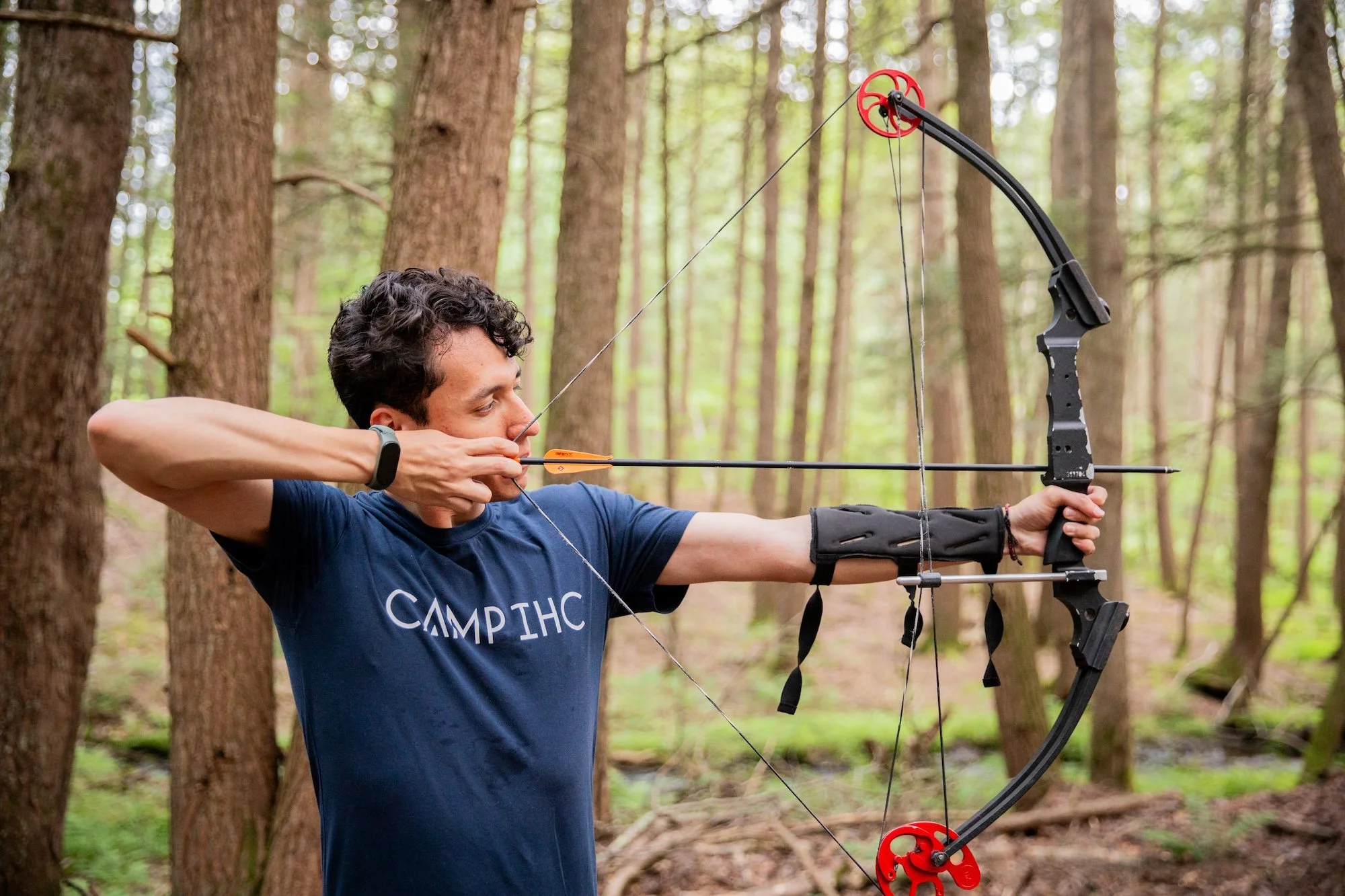 A man in a navy t-shirt aiming a recurve bow in a forest.