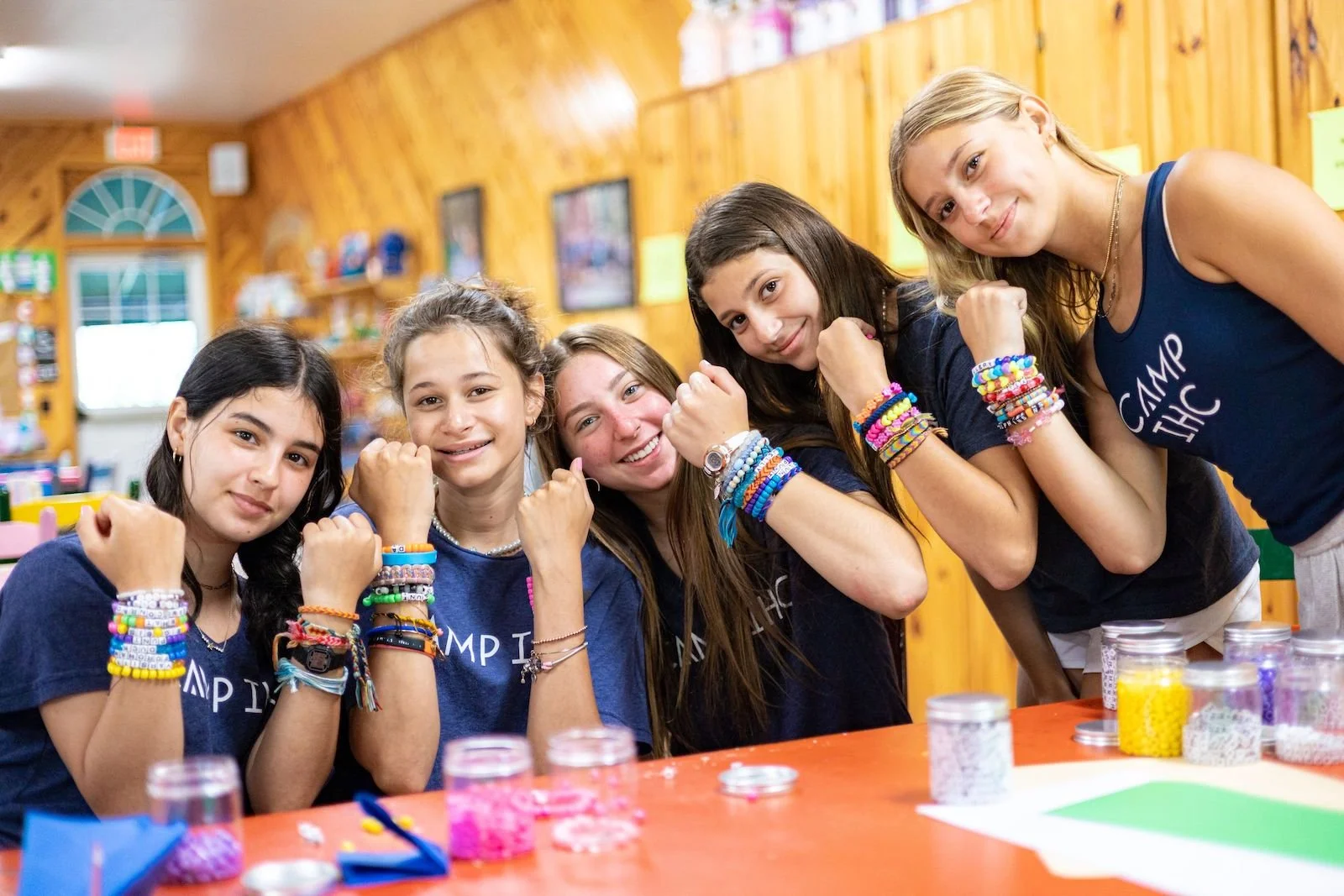 Group of five young girls at summer camp showing their muscular arms, wearing colorful friendship bracelets, sitting at a table with craft supplies in a wooden cabin.