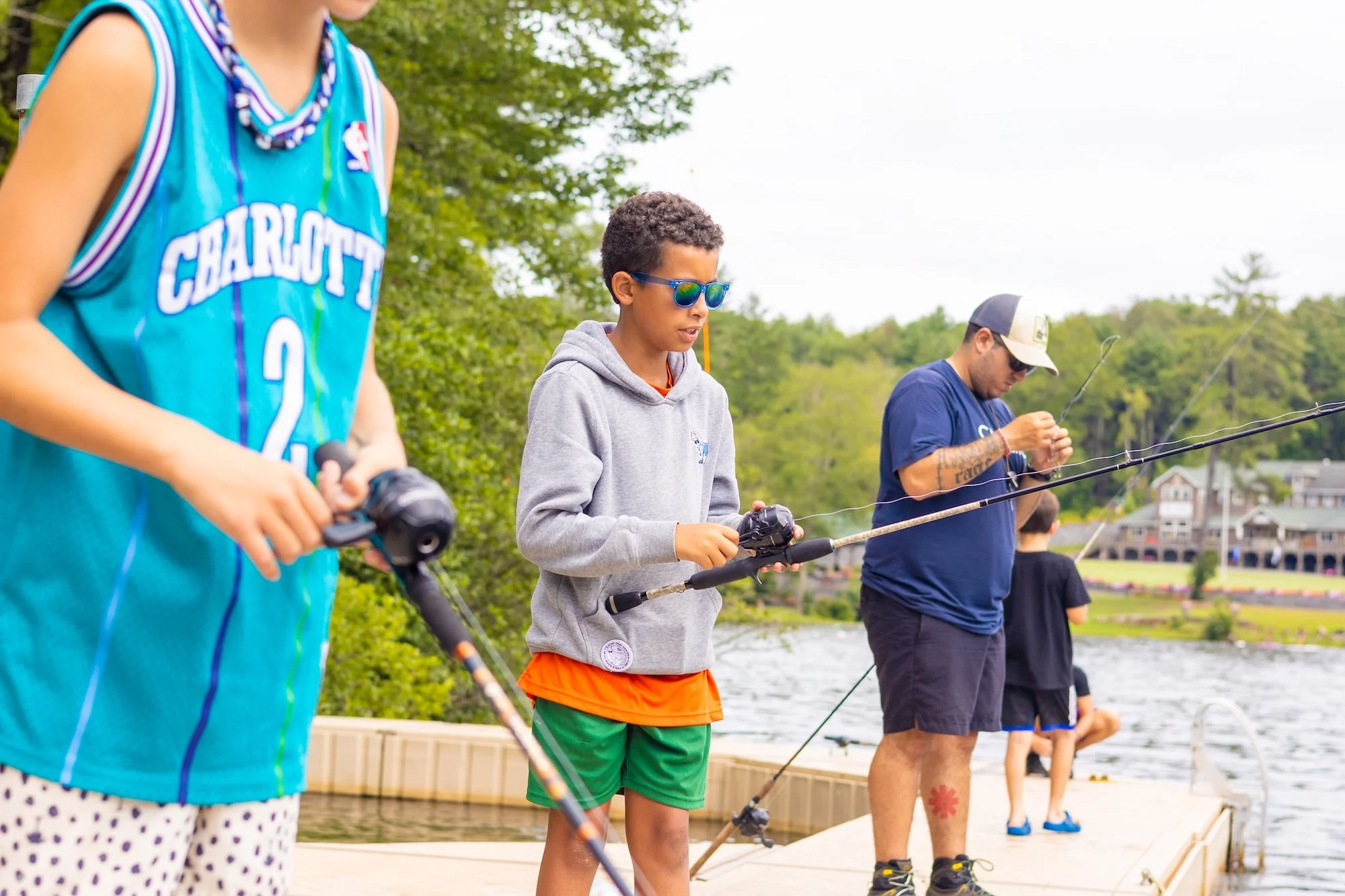 Three people fishing on a dock by a lake with trees in the background.
