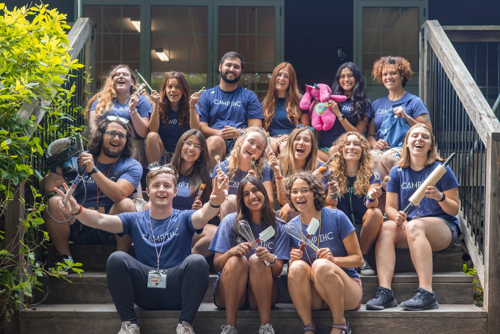 A group of young people wearing matching blue Camp IHC shirts sitting on the steps outside a building, smiling and holding various playful props like paintbrushes, a roller, whisk, and stuffed pink unicorn. They appear cheerful and excited, enjoying a camp activity or team photo.