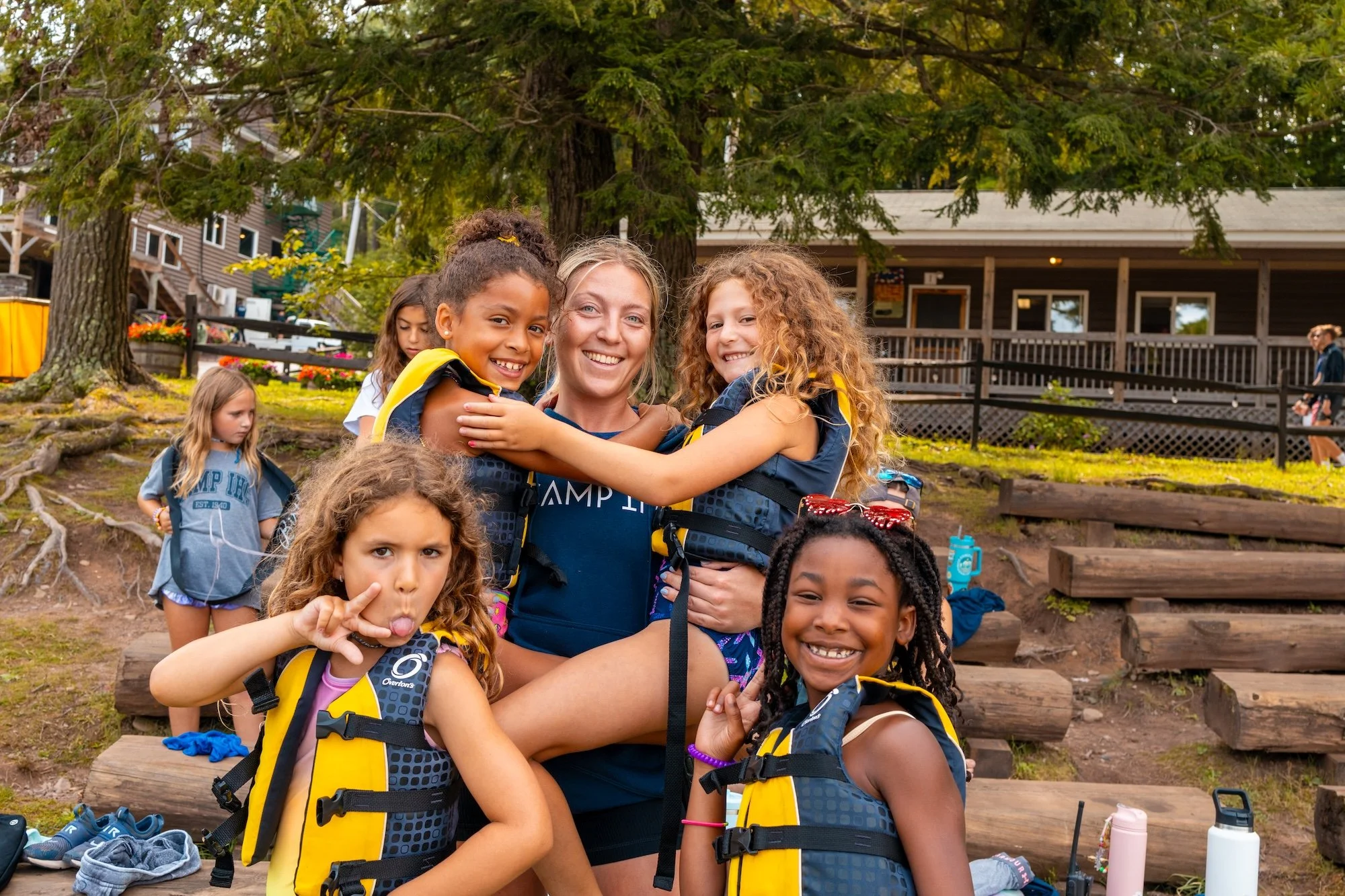 A group of young girls and a camp counselor posing outdoors, surrounded by trees and a wooden building, all wearing life jackets and smiling at the camera.