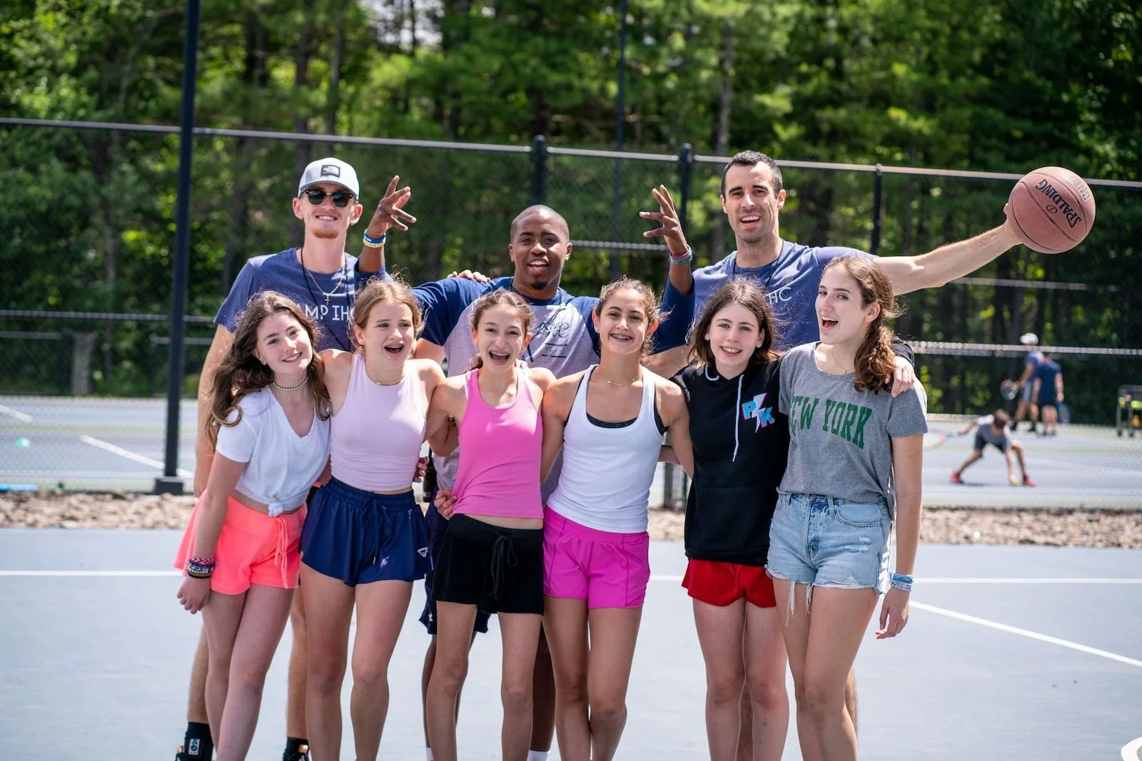 Group of smiling young female and male teenagers standing together on an outdoor tennis court, with some kids playing tennis in the background, surrounded by trees.