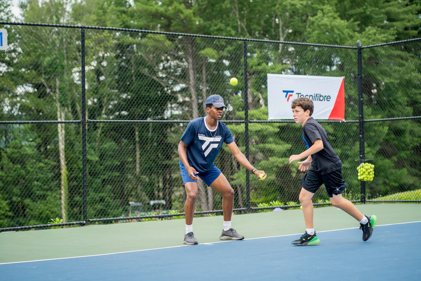 Two boys playing tennis on an outdoor court, one holding a tennis ball, with a chain-link fence and trees in the background.