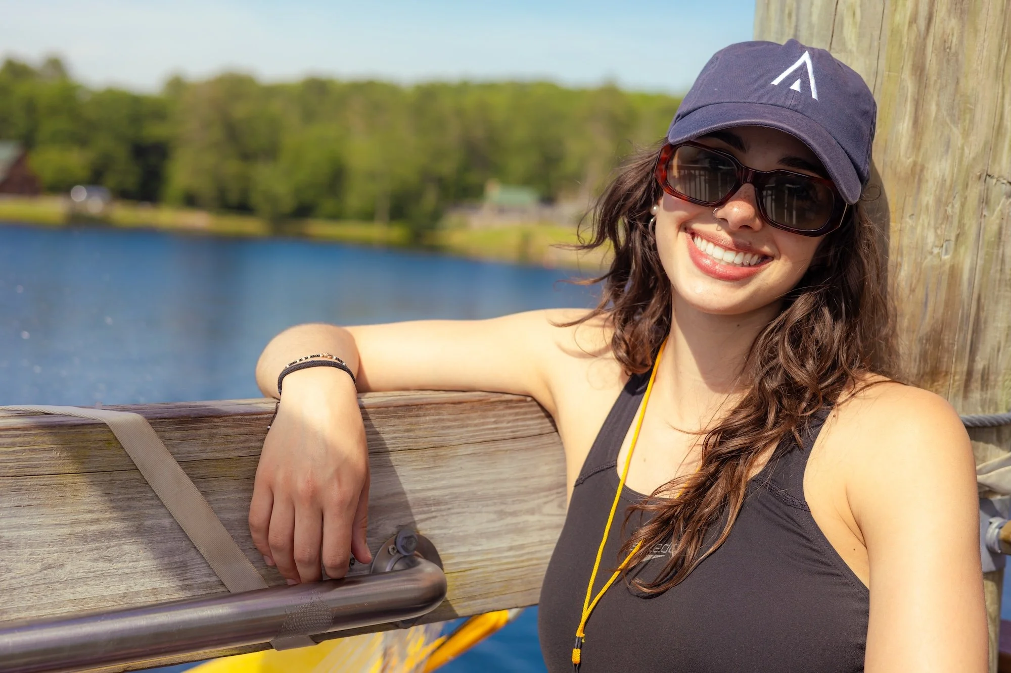 Young woman with curly brown hair wearing sunglasses, a navy cap, and a sleeveless black top, smiling and leaning on a wooden railing near a body of water during sunny day.