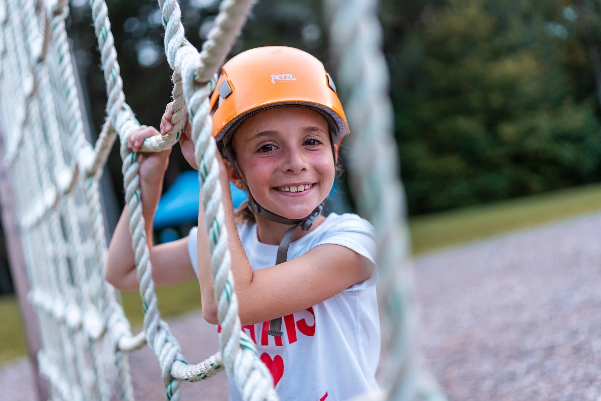 A smiling young girl wearing an orange helmet climbing on a rope structure at an outdoor playground.