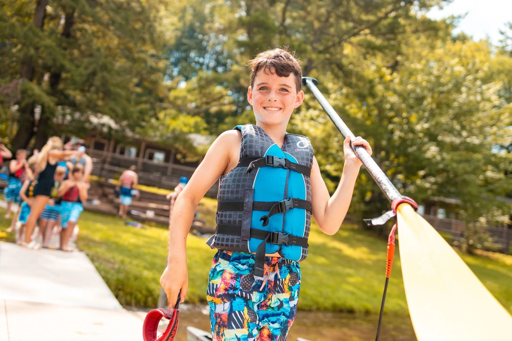 A smiling boy in a life jacket holding a water sports paddle, with a group of children in the background near a lake on a sunny day.