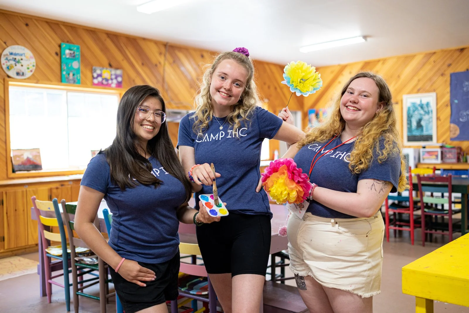 Three young women in blue T-shirts smiling inside a colorful room, holding yellow and pink paper flowers, with tables and chairs in the background.