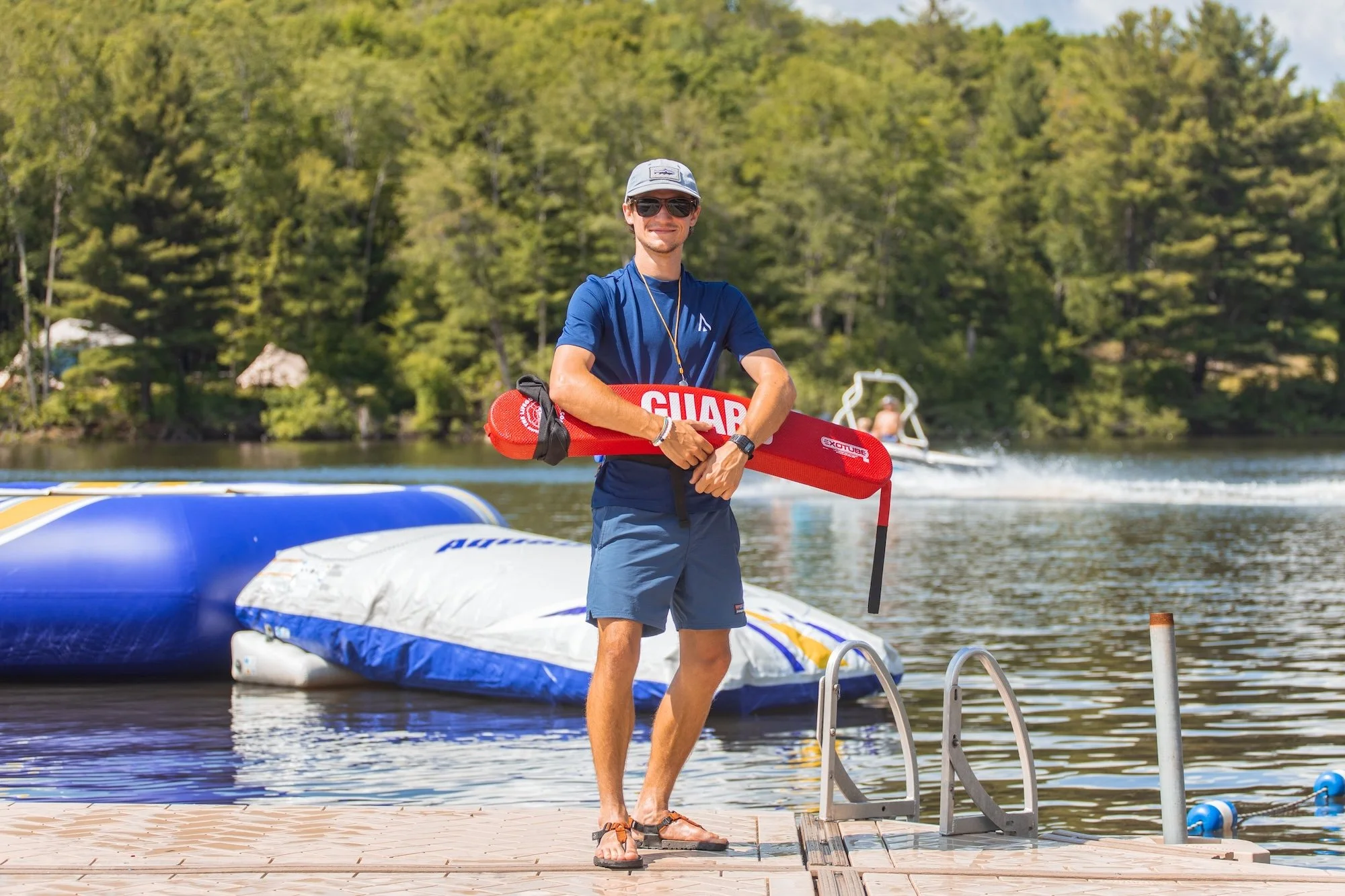 A man in a blue t-shirt, shorts, and sunglasses standing on a dock at a lake, holding a red rescue buoy, with inflatable water toys and a person on a boat in the background.