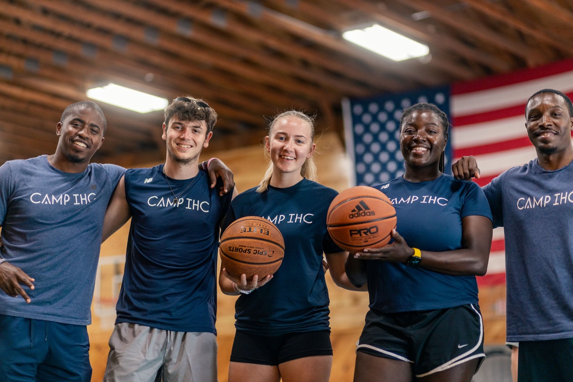 Group of five smiling young people standing together in a gymnasium, some holding basketballs, with an American flag hanging in the background.