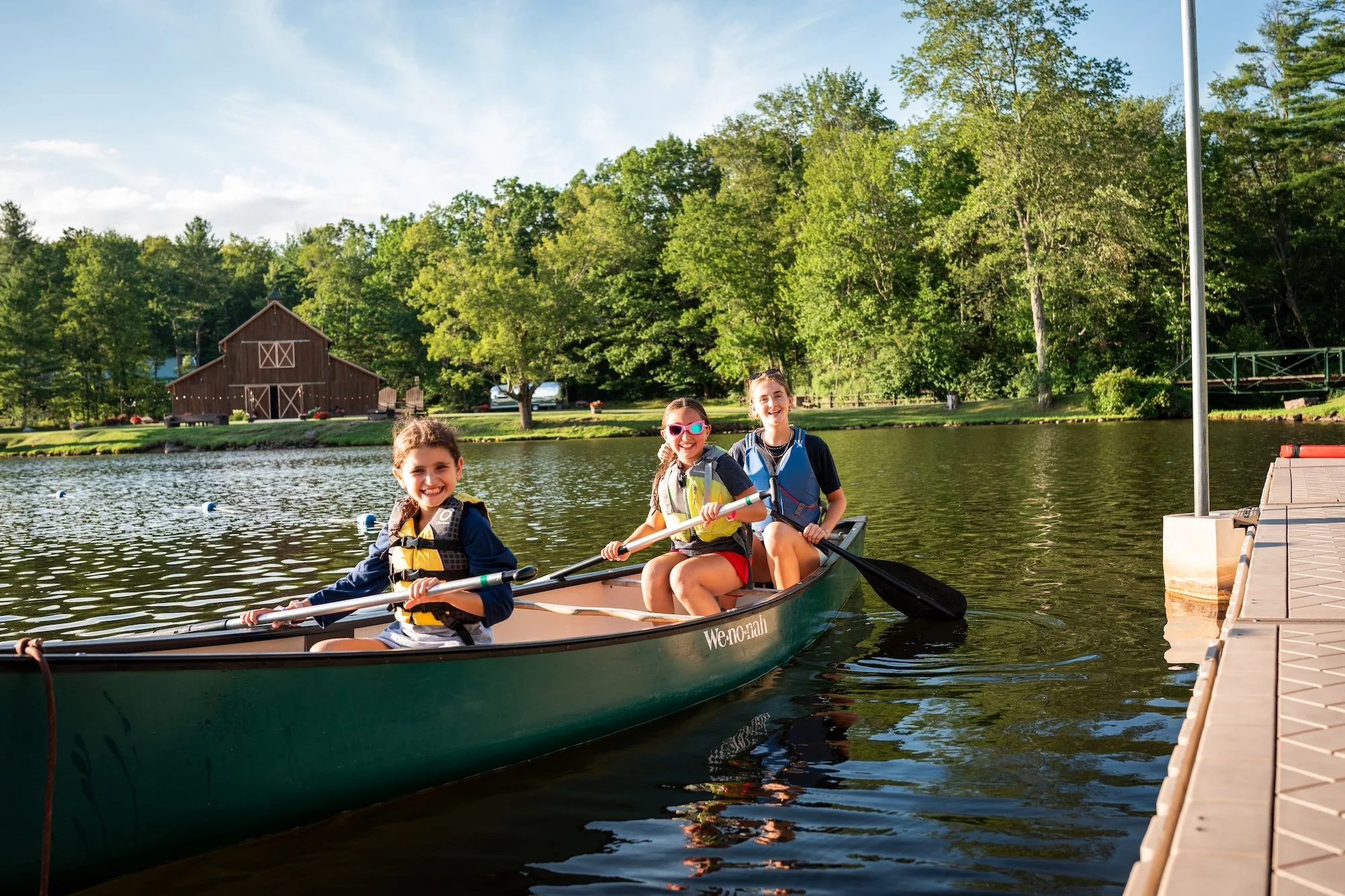 Three children in a green canoe on a lake. They are smiling and paddling with black oars, surrounded by trees and a wooden boat house in the background.