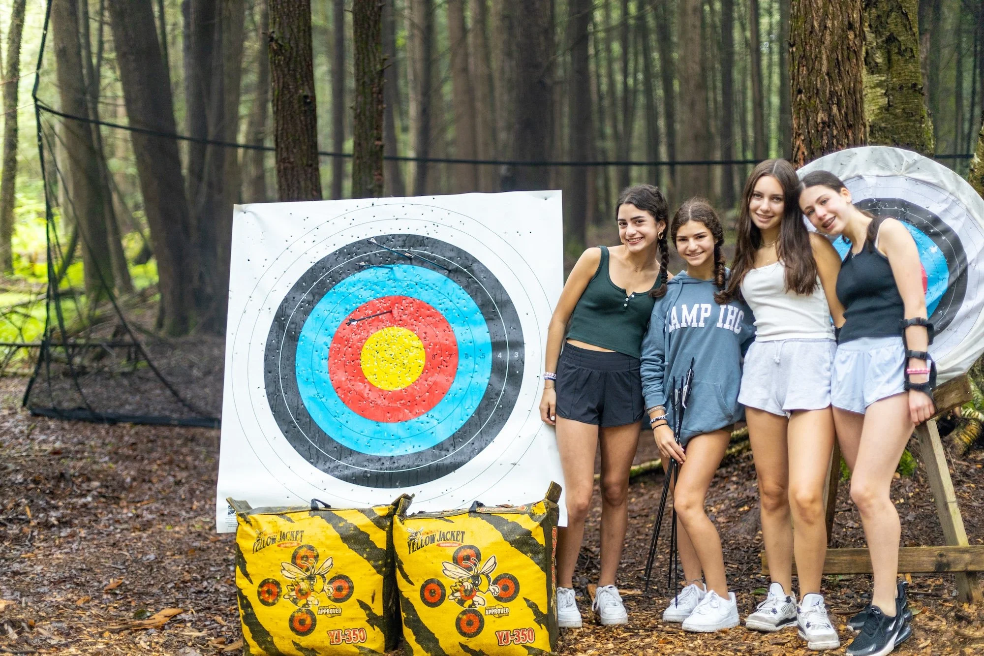 Four young women standing in a wooded area after archery practice, with a large target and quiver bags in front of them.